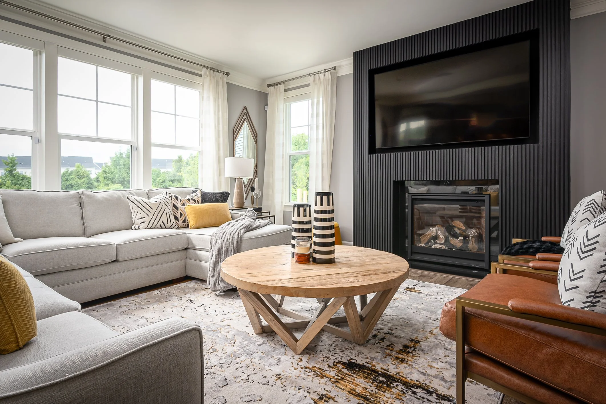 Modern living room with large windows, a light gray sofa, a round wooden coffee table with striped vases, a fireplace with a black textured wall, a wall-mounted TV, decorative pillows, and a patterned area rug.