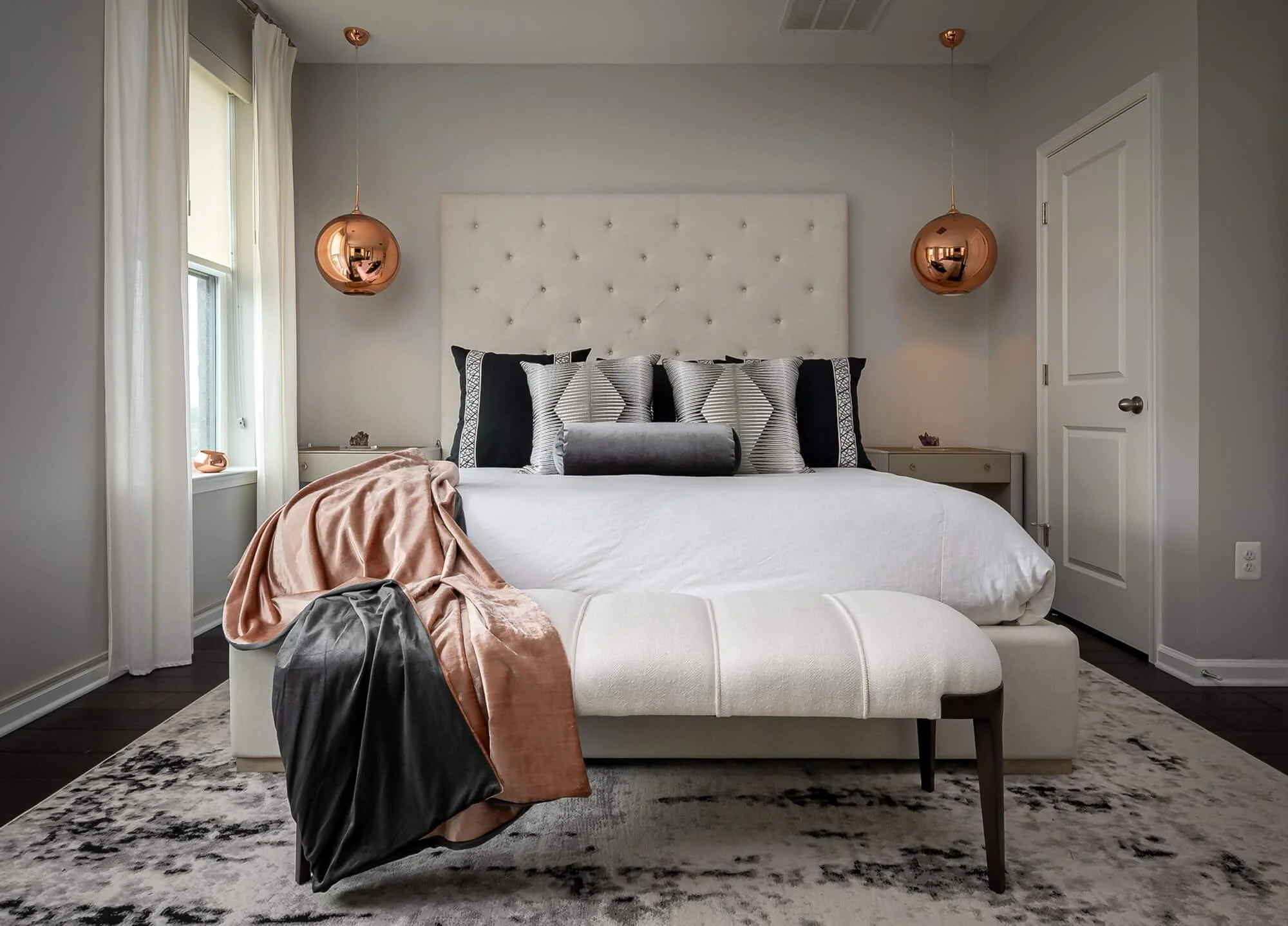 A modern bedroom with a large white bed, decorated with black, gray, and striped pillows, a gray velvet bolster, and a soft pink blanket. Flanking the bed are two white nightstands with small decorative items. Above the bed, two copper pendant lights