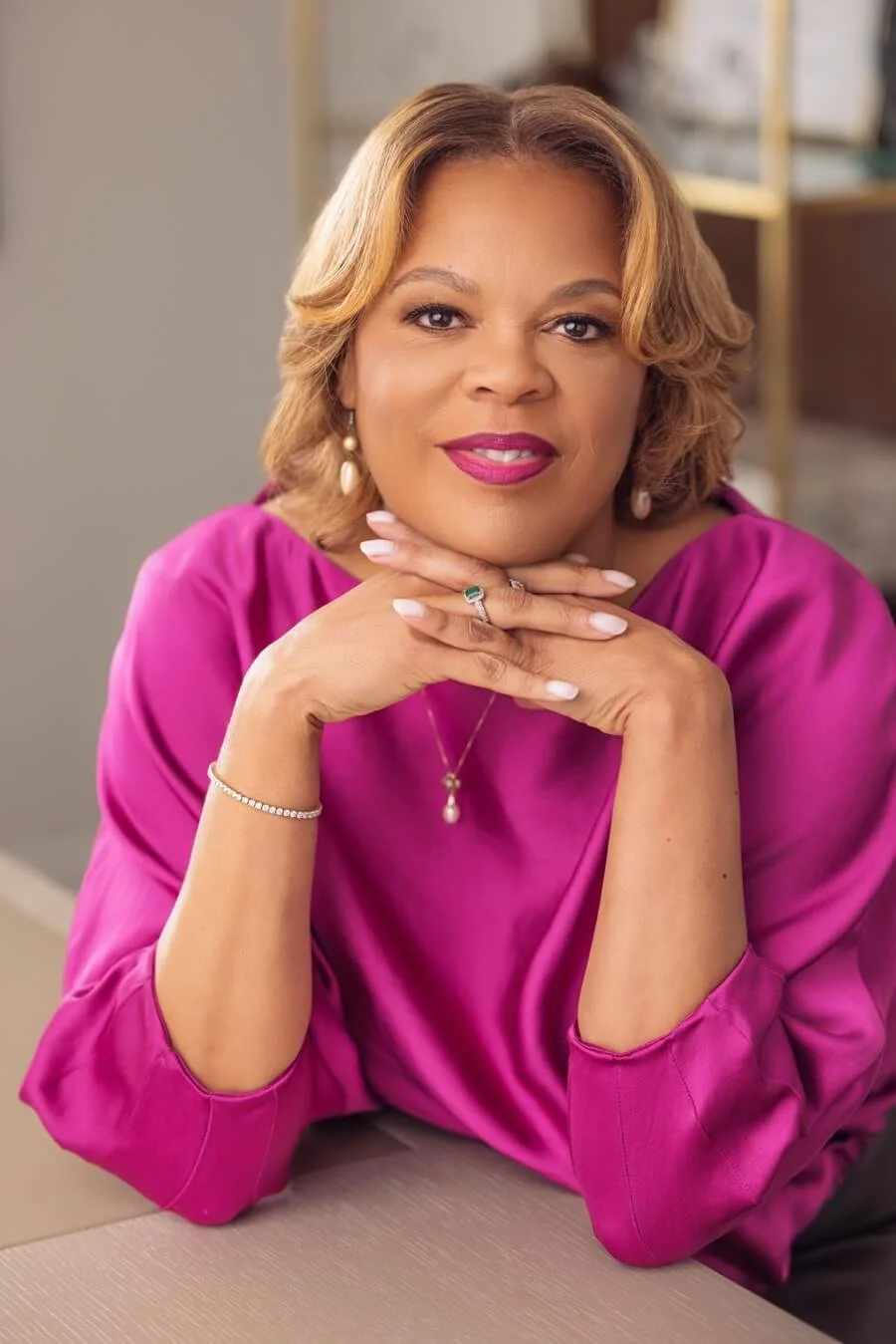 Monique Holland, wearing a pink blouse, earrings, and jewelry, sitting with her hands under her chin, smiling at the camera.