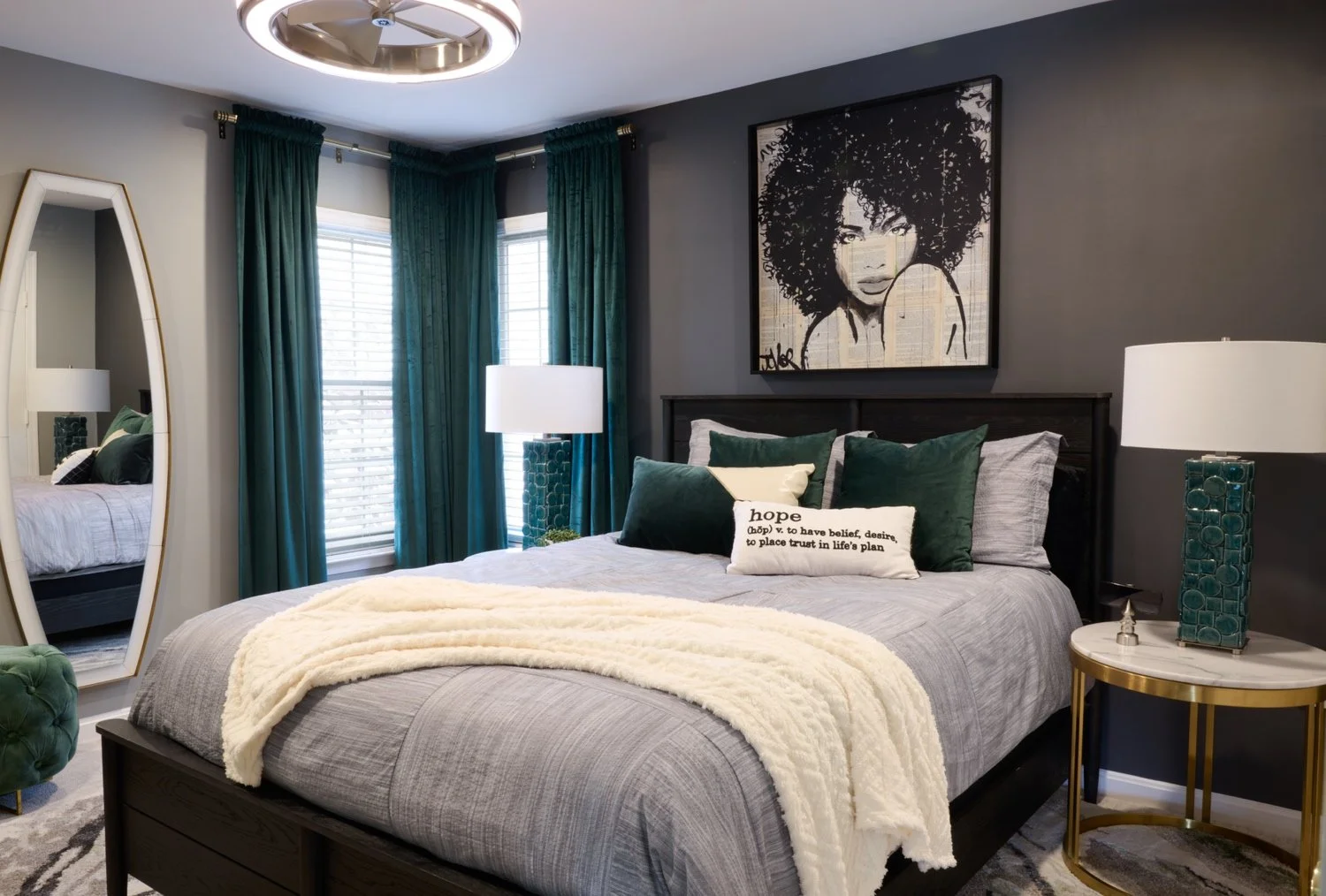 A bedroom with a dark wood bed, gray bedding, and several green pillows. A white pillow with the word pink hope is on the bed. There are teal curtains and two lamps with teal bases on nightstands. A large portrait of a woman with curly hair hangs above the bed, and a tall, oval mirror is on the left side of the room.