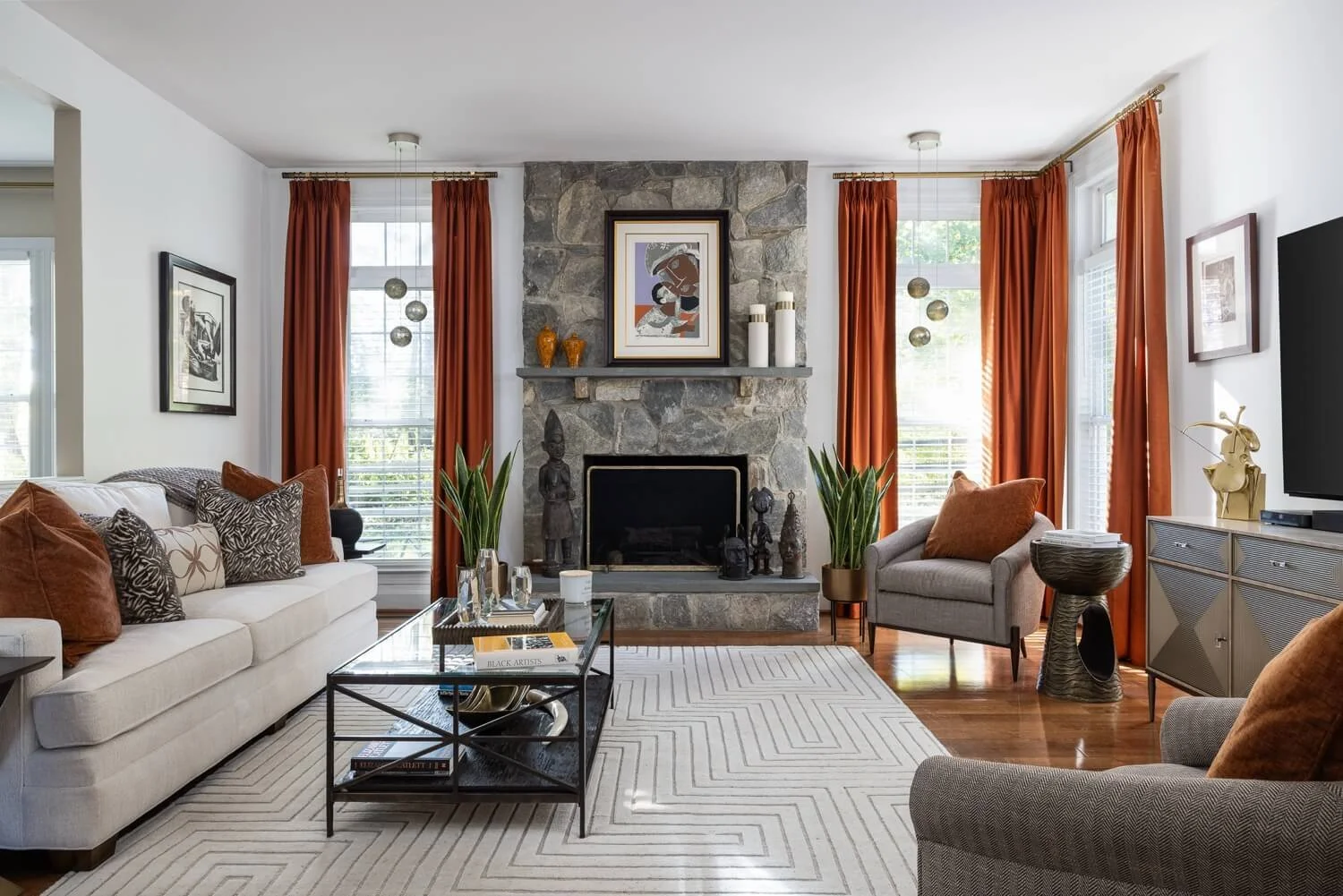 Living room created by Monique with white sofa, gray armchair, and orange curtains, centered around a stone fireplace, decorated with artwork, sculptures, and plants, and a glass coffee table on a white patterned rug.