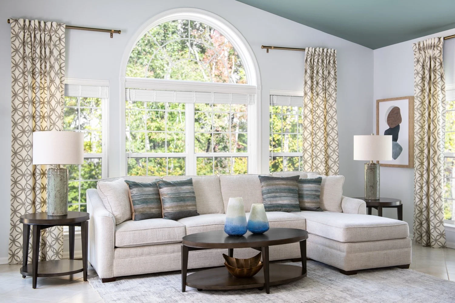 Living room with large arched window, beige sectional sofa with patterned pillows, dark wood coffee table with two blue to white gradient vases, two side tables with lamps, patterned curtains, and wall art, with trees visible outside.