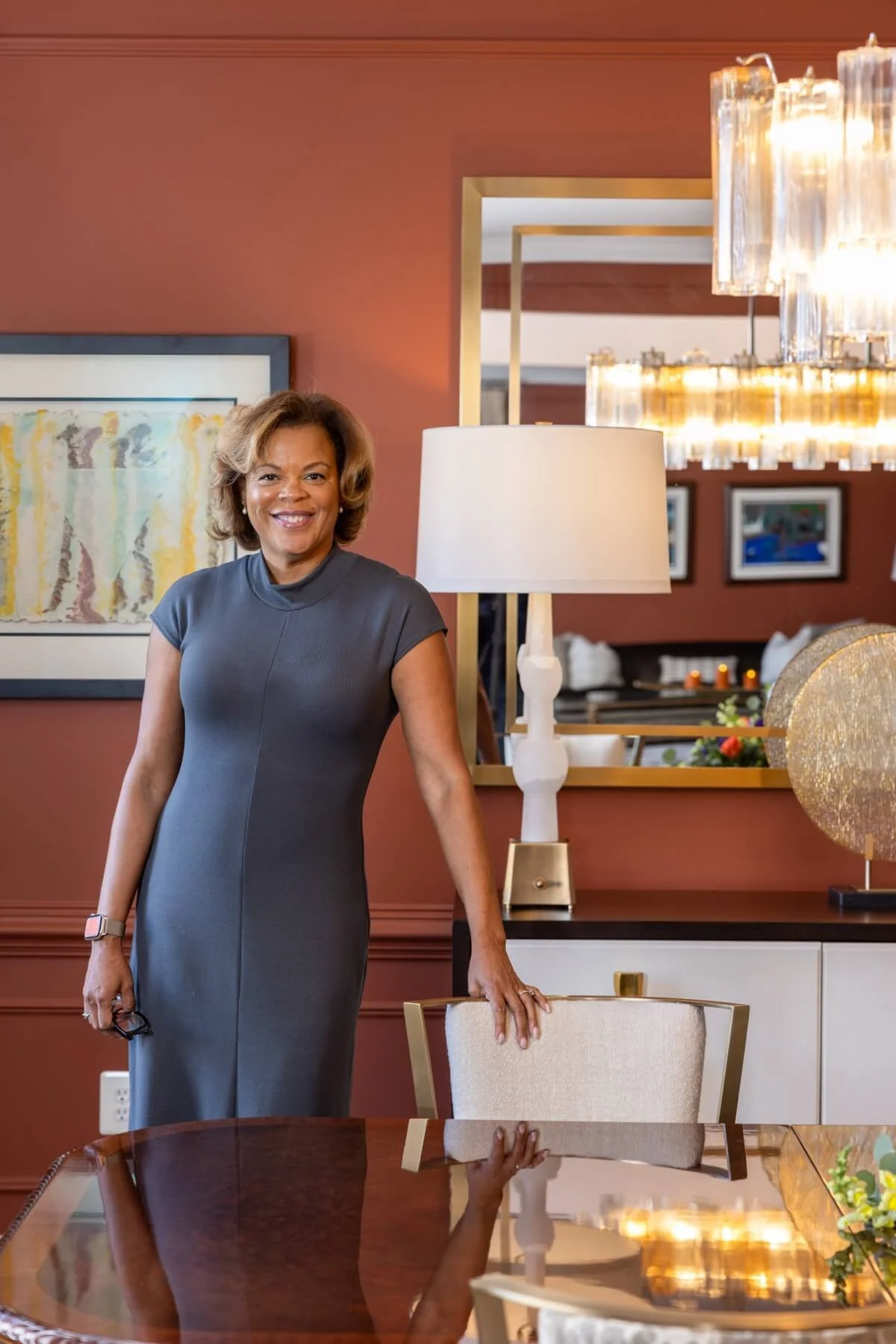 Monique in a gray dress smiling and standing in a dining room with a polished wooden table, a beige upholstered chair, a white table lamp, framed artwork, and decorative lighting fixtures.