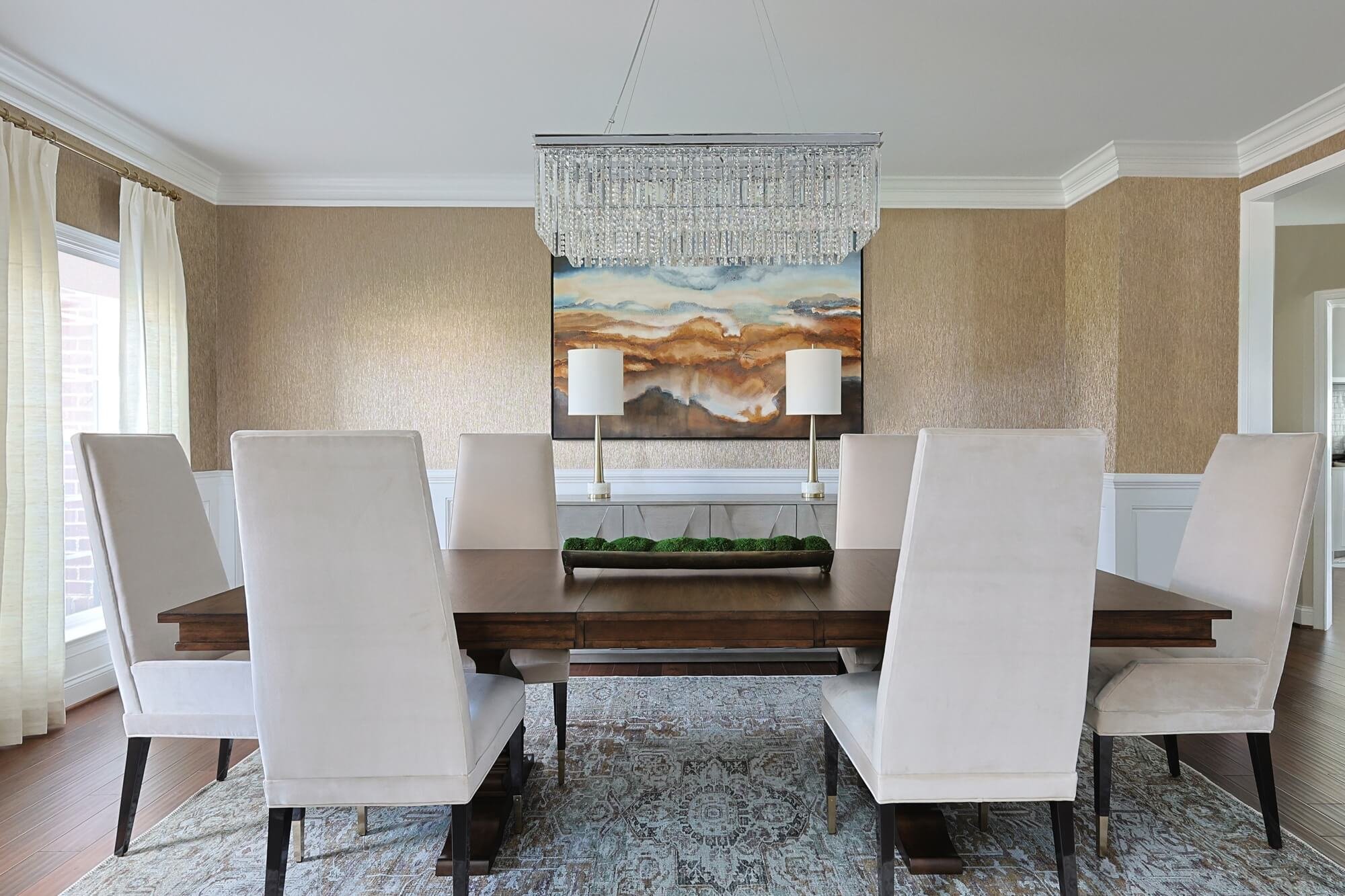 A dining room with seven white high-back chairs around a wooden table, two white table lamps, a large abstract landscape painting on the wall, a crystal chandelier hanging above, and light-colored curtains near a window.