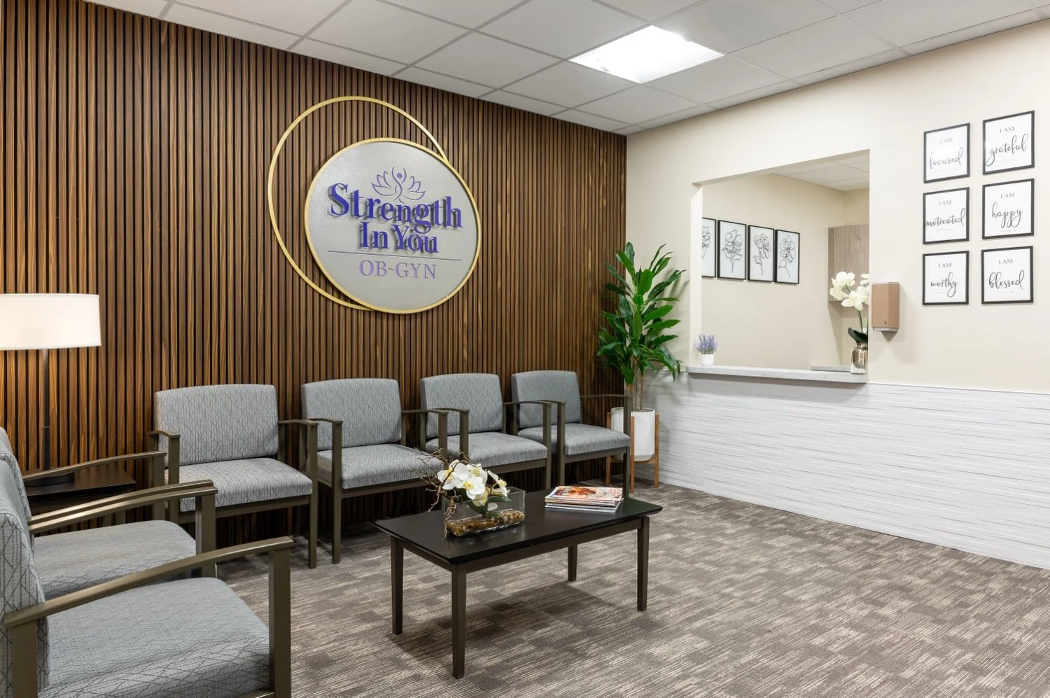 Waiting area in a medical office with a brown wooden wall featuring a sign that reads "Strength In You OB-GYN," four gray chairs, a black coffee table with magazines and a flower arrangement, green potted plant, and framed quotes on the walls.