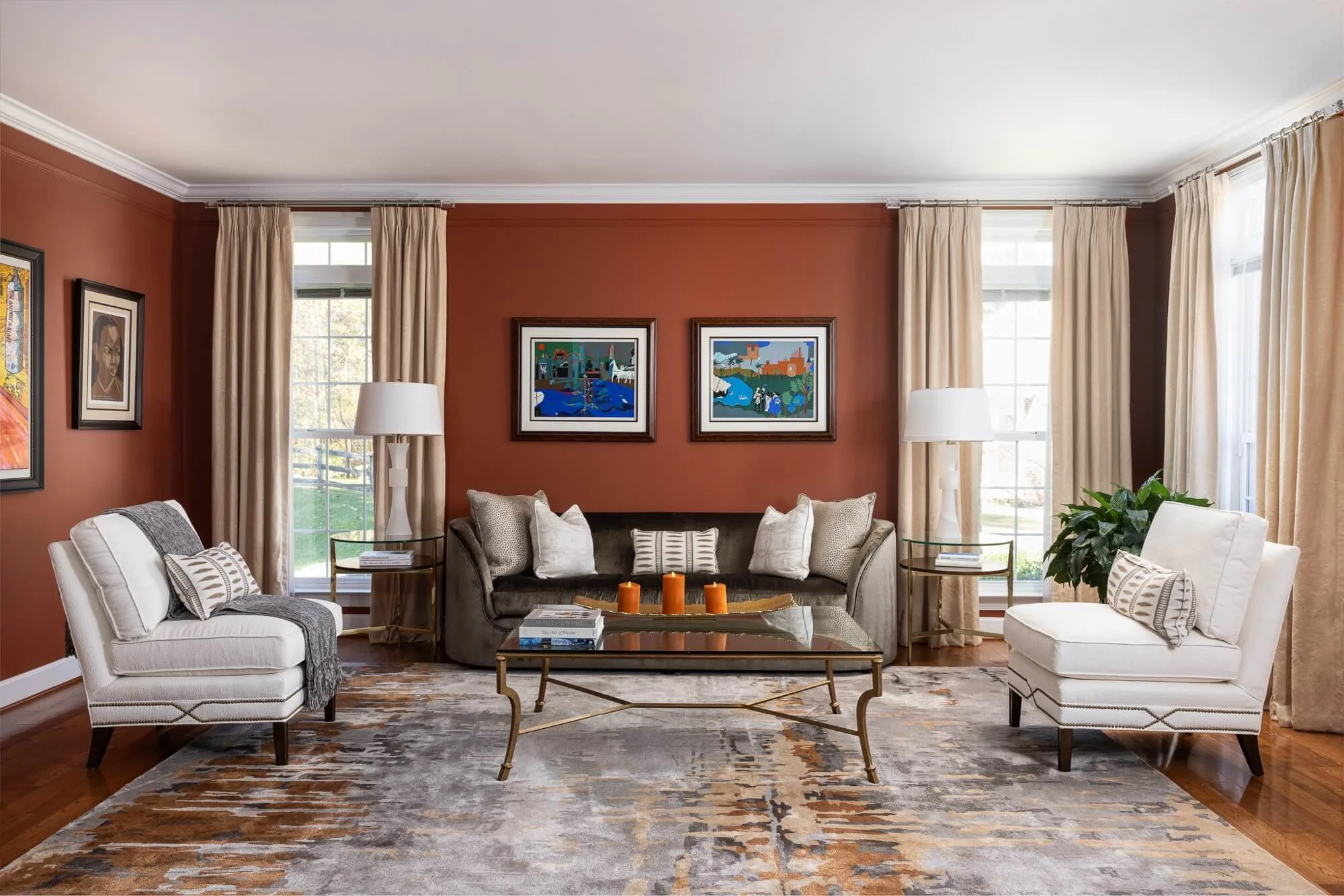 Living room with a brown accent wall, beige curtains, two white armchairs, a gray sofa, a glass coffee table, paintings on the wall, and large windows.