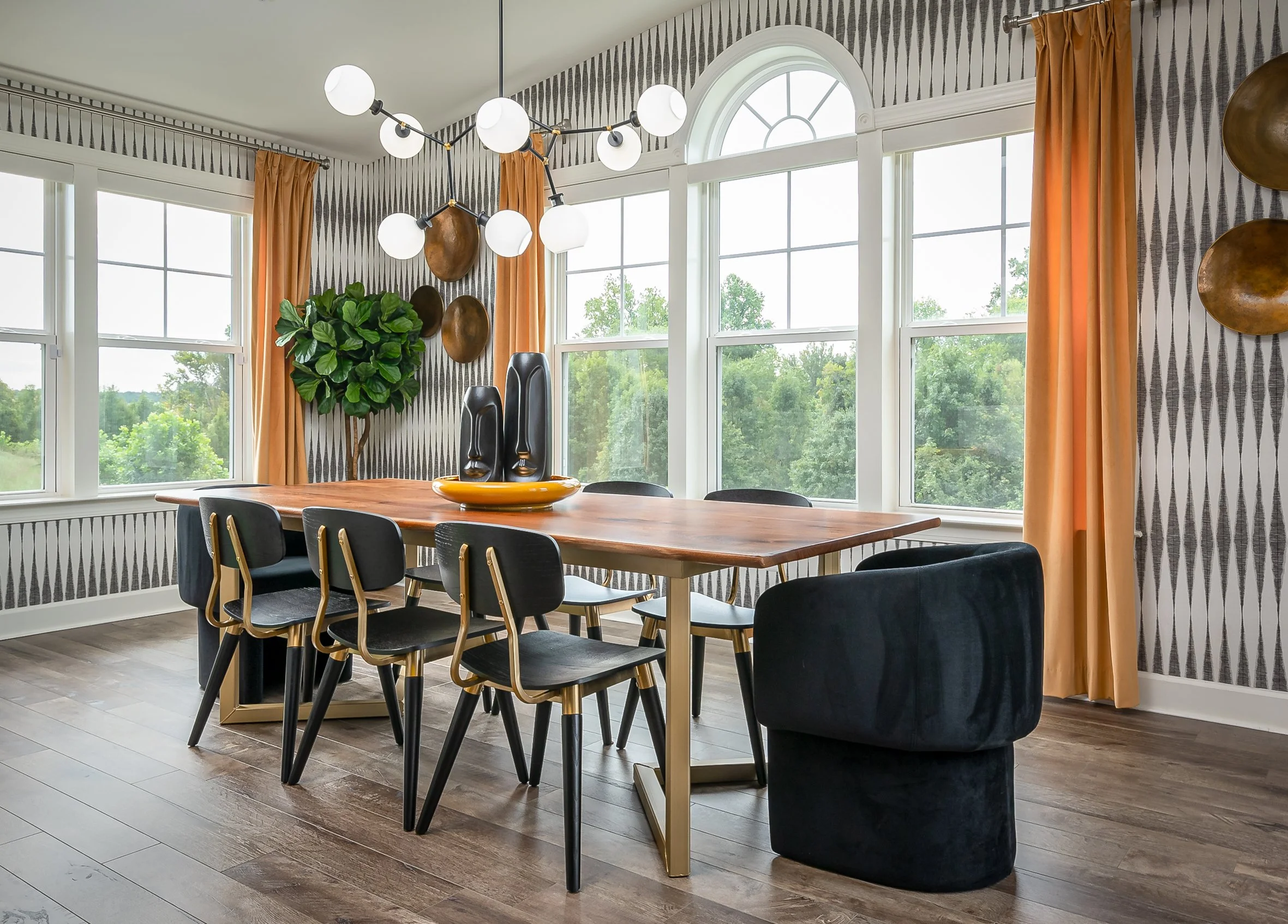 Modern dining room with large windows, wooden table, black and gold chairs, orange curtains, geometric wallpaper, wall decor, and a chandelier with white globes.