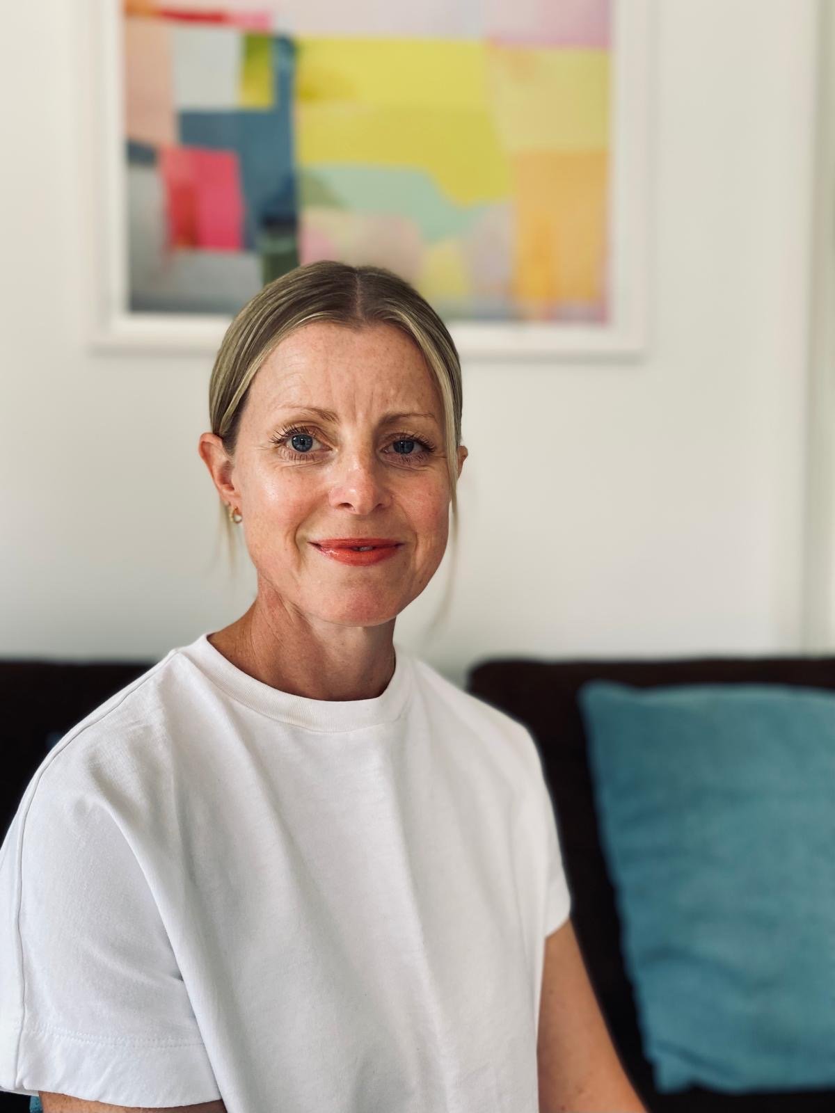 A woman with blonde hair and blue eyes, wearing a white t-shirt, sitting indoors with a colorful abstract painting on the wall behind her.