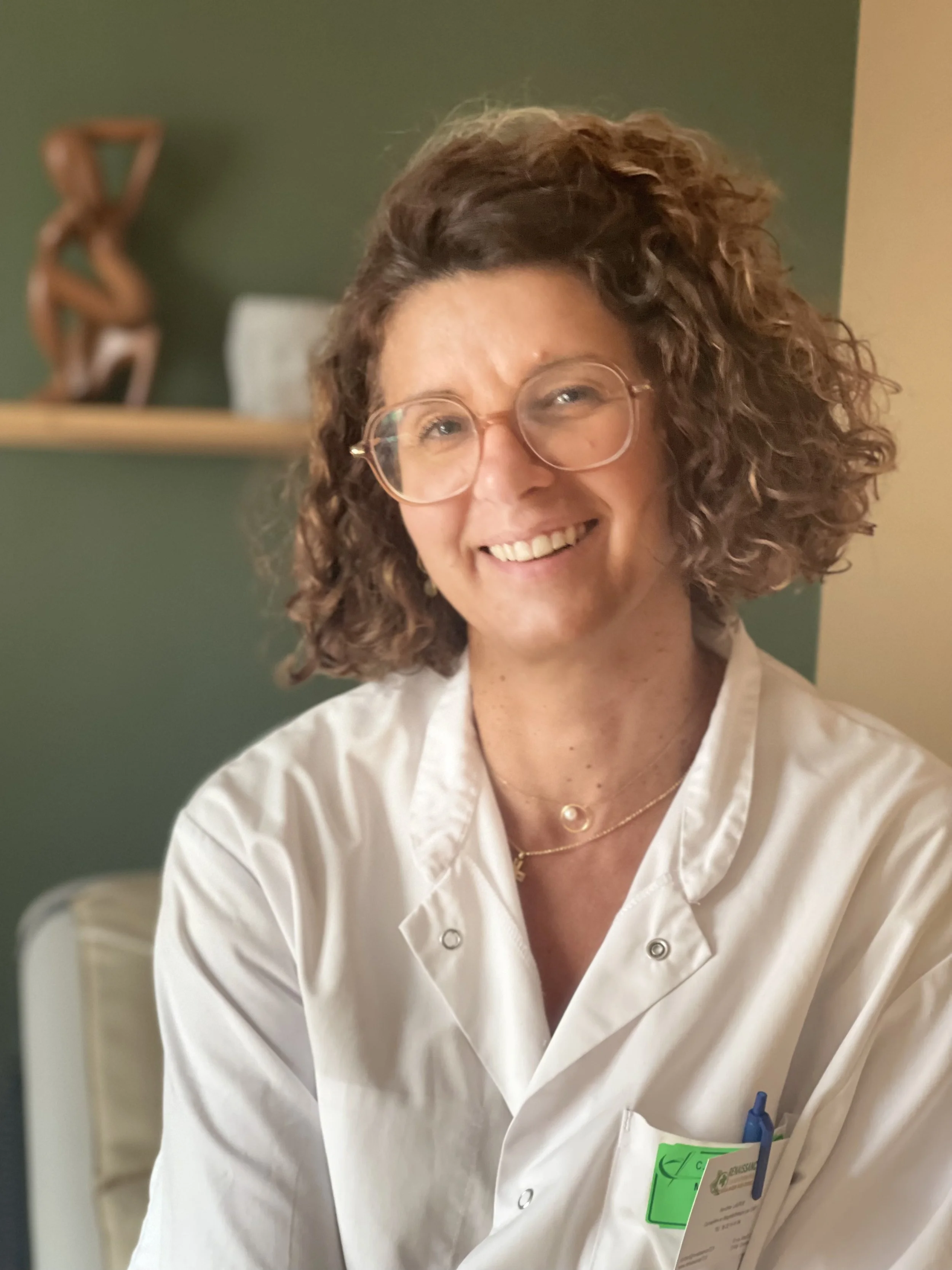 Une femme souriante portant une blouse blanche et des lunettes, assise dans un intérieur avec un mur vert et une étagère en arrière-plan.