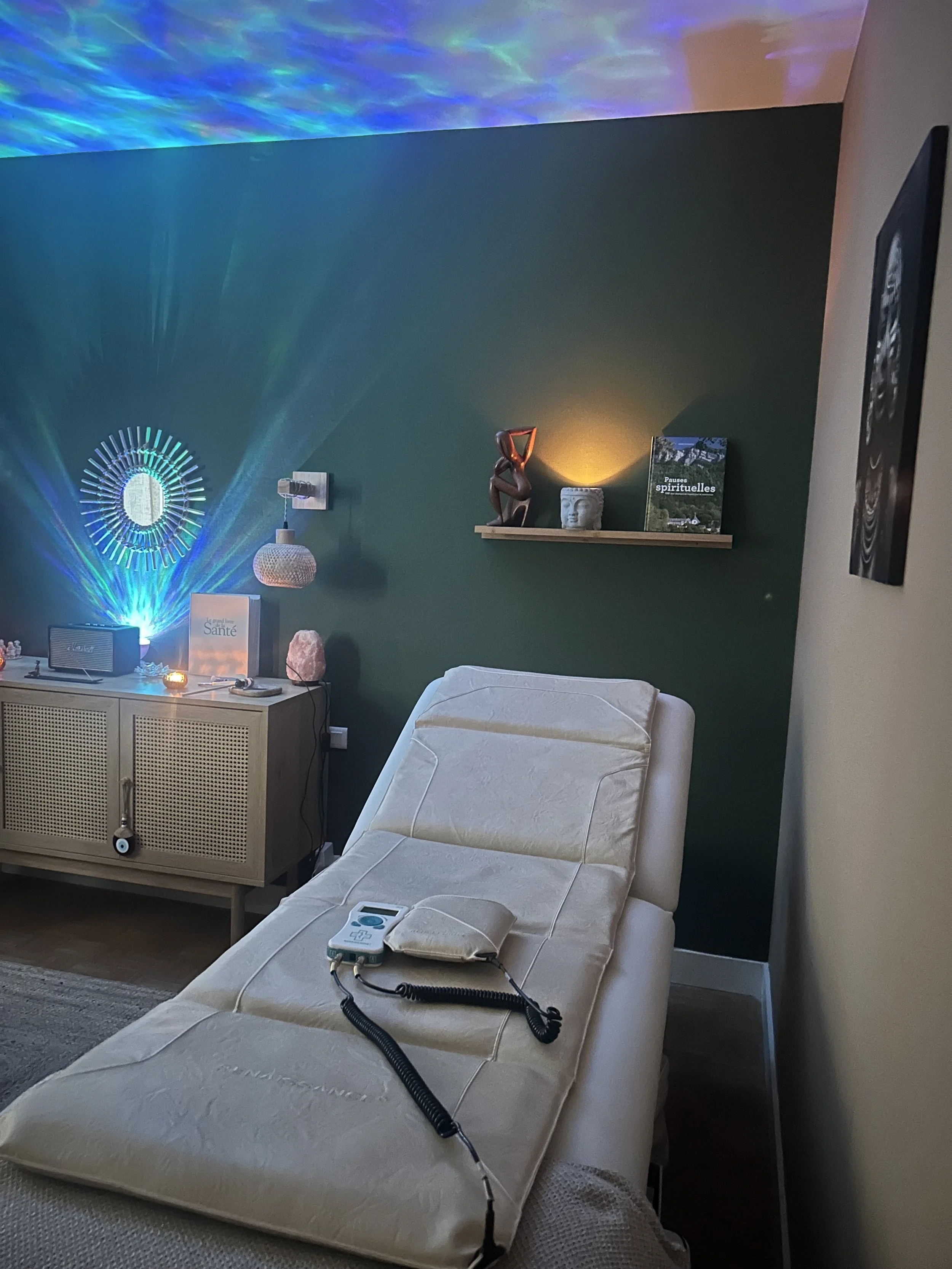 Une salle de soins ou de relaxation avec un lit inclinable, une décoration d'ambiance avec des lumières colorées, des étagères avec des objets décoratifs, un petit meuble avec des livres et une enceinte, et un éclairage tamisé.