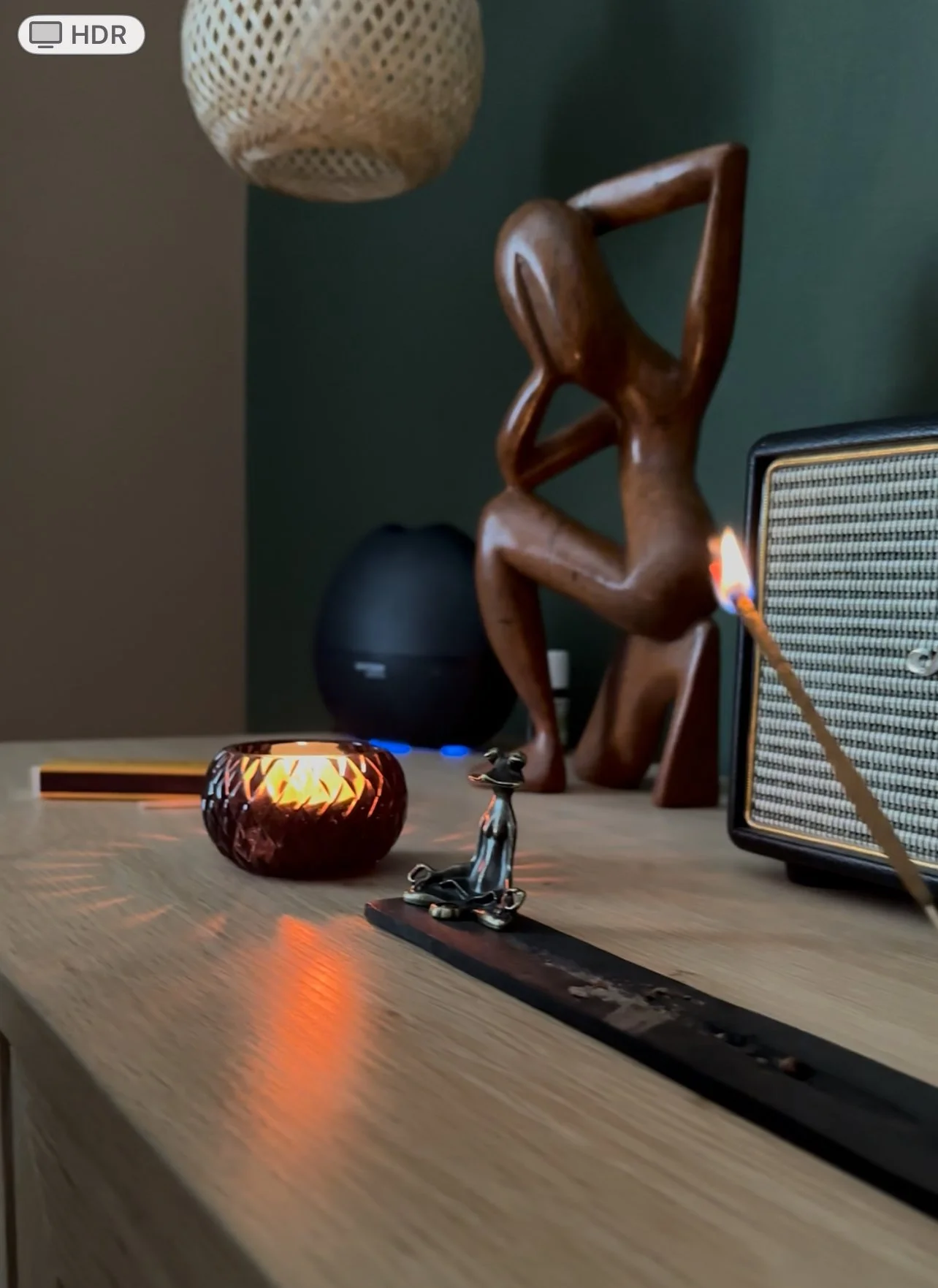 Une collection d'objets décoratifs sur une surface en bois, comprenant une figurine en métal d’un chien, une bougie allumée, une sculpture en bois d’une femme assise, un haut-parleur, et une lampe suspendue. En arrière-plan, un mur vert foncé.