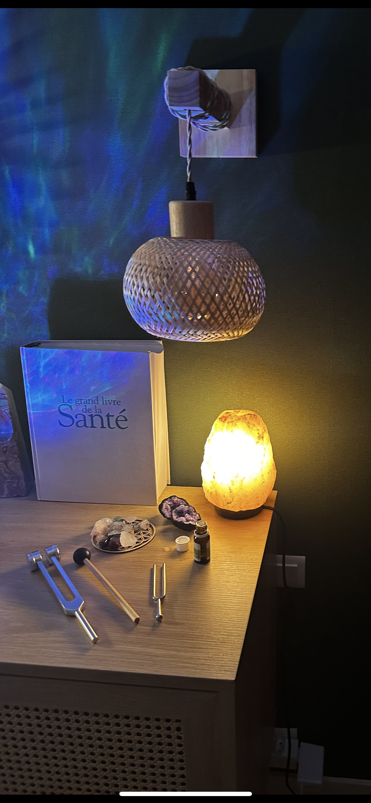Une table en bois avec un livre intitulé 'Le grand livre de la Santé', un lampadaire en pierre salée émettant une lumière chaude, un plateau avec des cristaux, une petite bouteille d'huile essentielle, et trois outils métalliques, avec une lampe suspendue en haut, le tout dans une pièce avec un mur vert foncé.