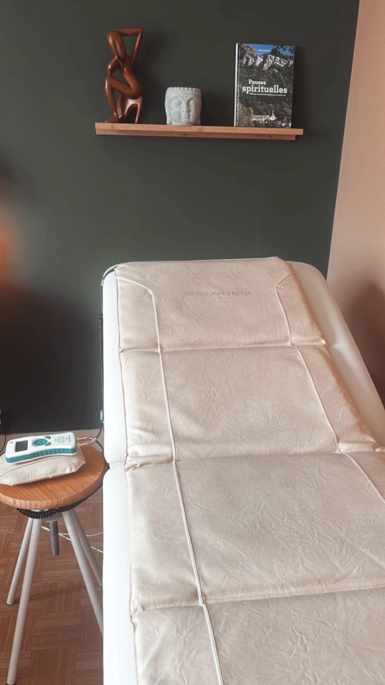 Une salle de soins ou de consultation avec un lit d'examen beige, une petite table en bois avec un appareil médical ou un tensiomètre, un mur vert foncé avec une étagère en bois contenant une sculpture en bois sujet de la tête humaine, un vase blanc en forme de visage, et un livre intitulé "Pauses spirituelles".
