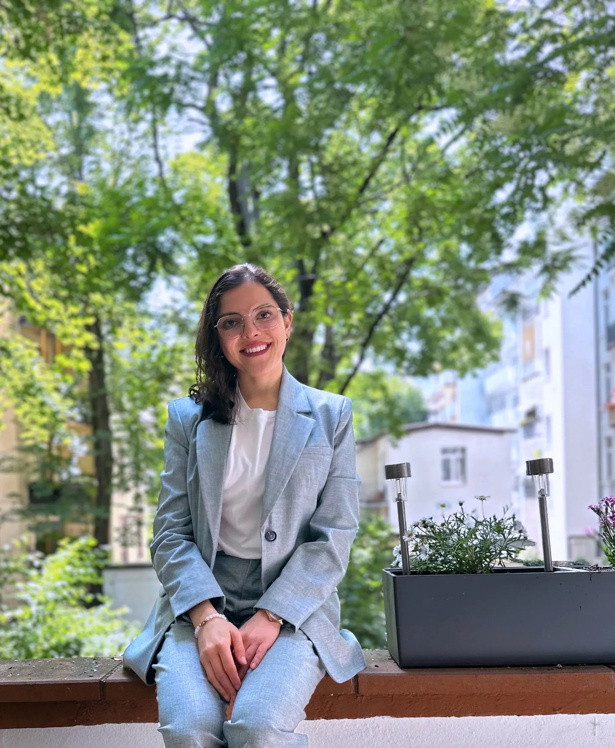 A woman in a light gray suit sitting on a balcony with greenery and trees in the background.