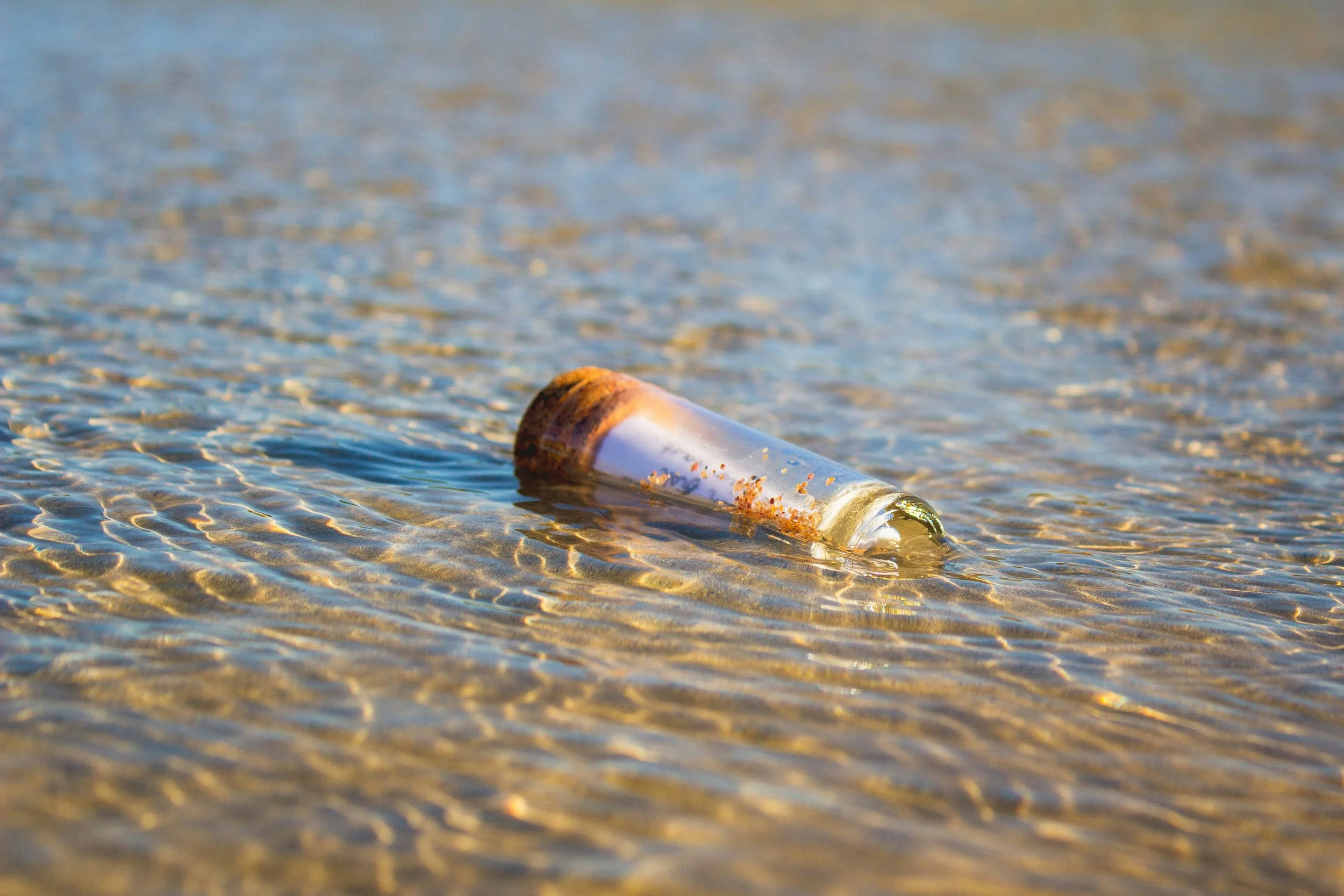 A glass bottle with a cork floating in shallow water on a sandy beach
