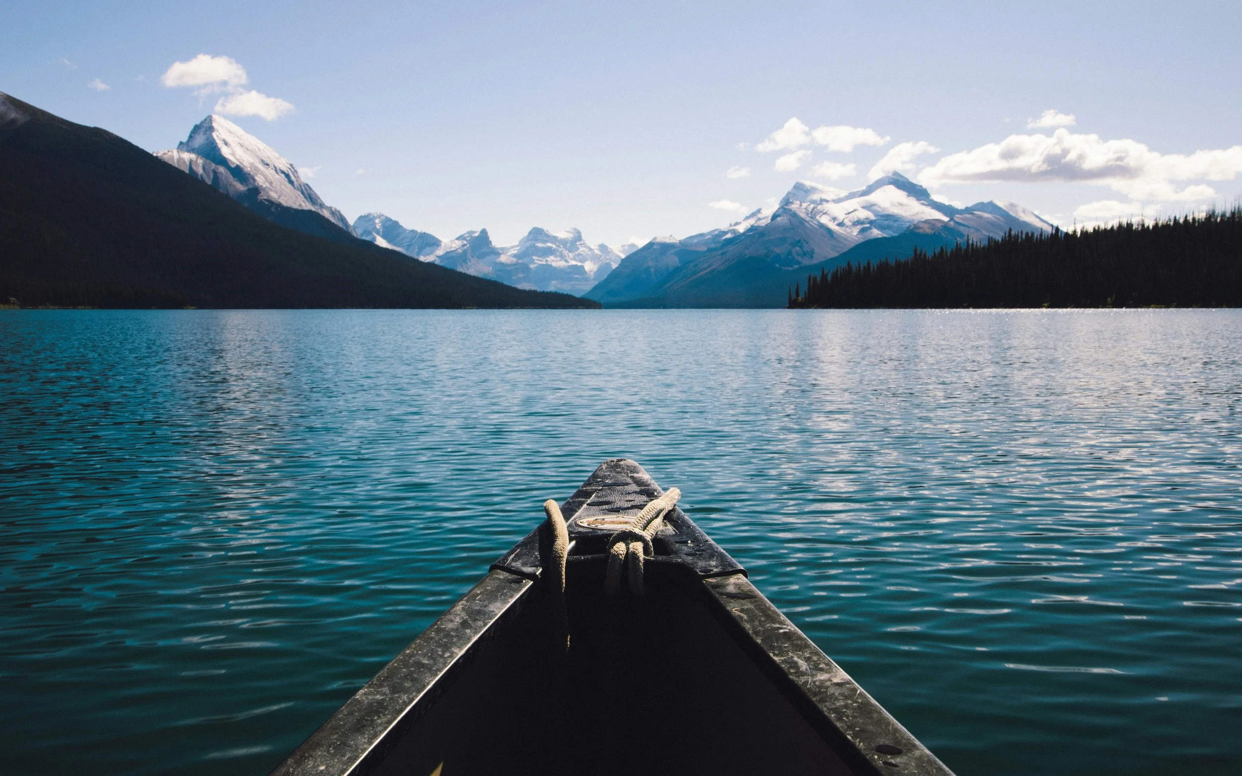 View from the front of a canoe on a large lake surrounded by mountains with snow caps, some trees along the shoreline, and partly cloudy sky.