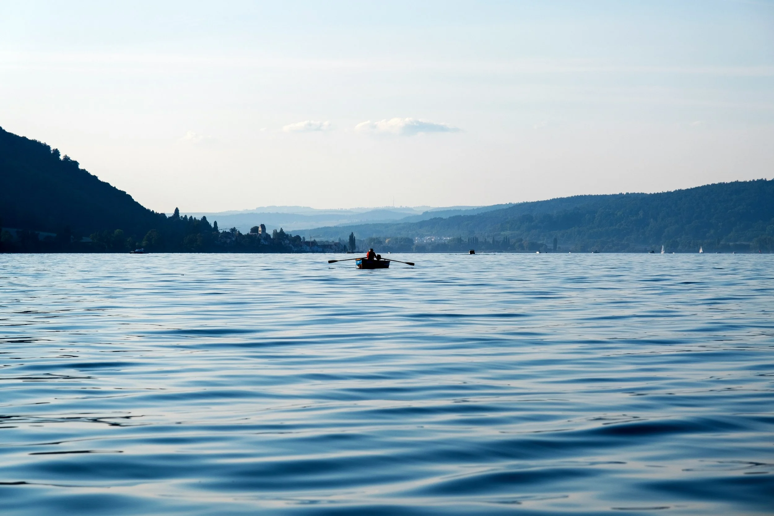 A person rowing a boat on calm lake with rolling hills in the background during daytime.