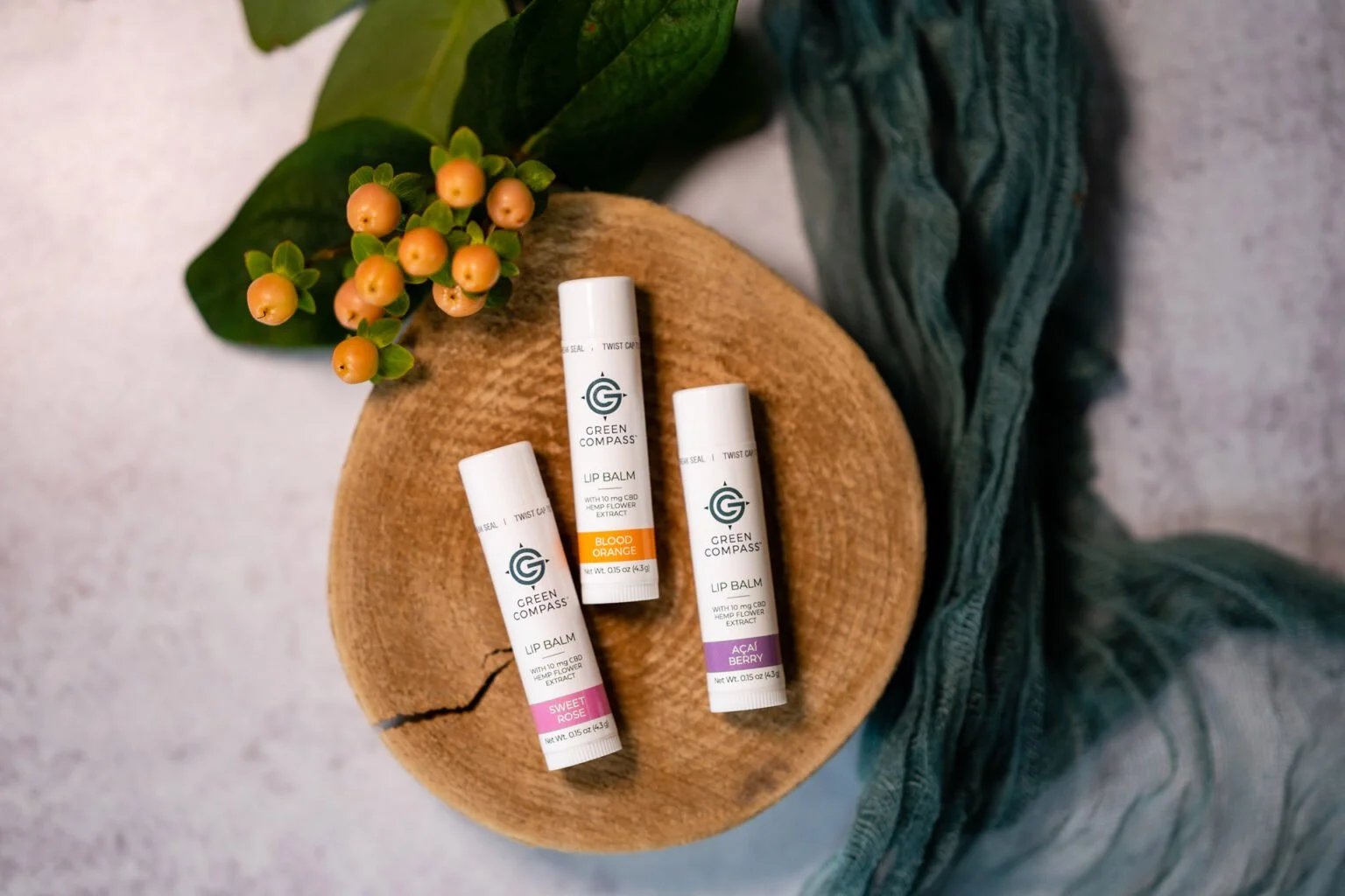 Green Compass lip balm tubes labeled Sweet, Blood Orange, and Acai Berry placed on a wooden slice, with a sprig of green leaves and orange berries nearby on a textured surface.