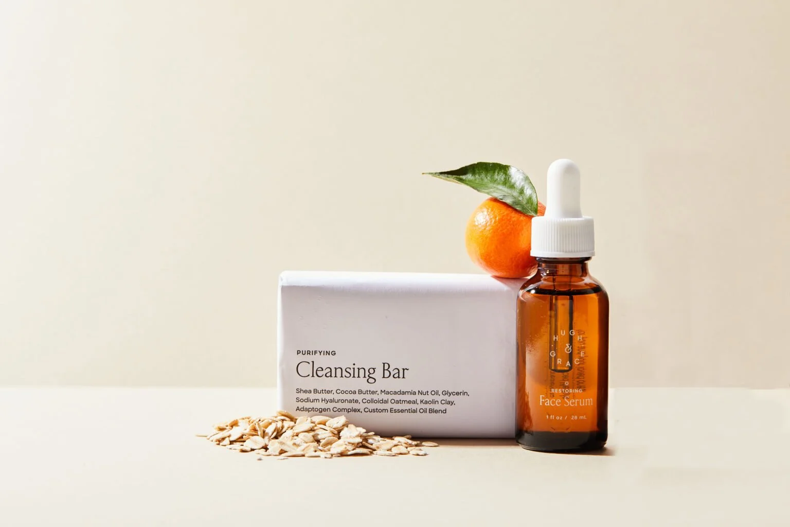 Hugh & Grace skincare product setup with a box of purifying cleansing bar, a glass bottle of face serum with a dropper, an orange with a green leaf on top, and sunflower seeds arranged on a light surface against a neutral background.