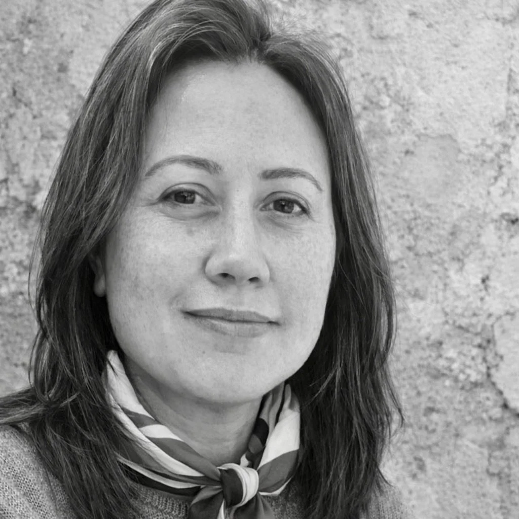 A woman with shoulder-length hair, wearing a scarf around her neck, smiling softly against a textured wall background.