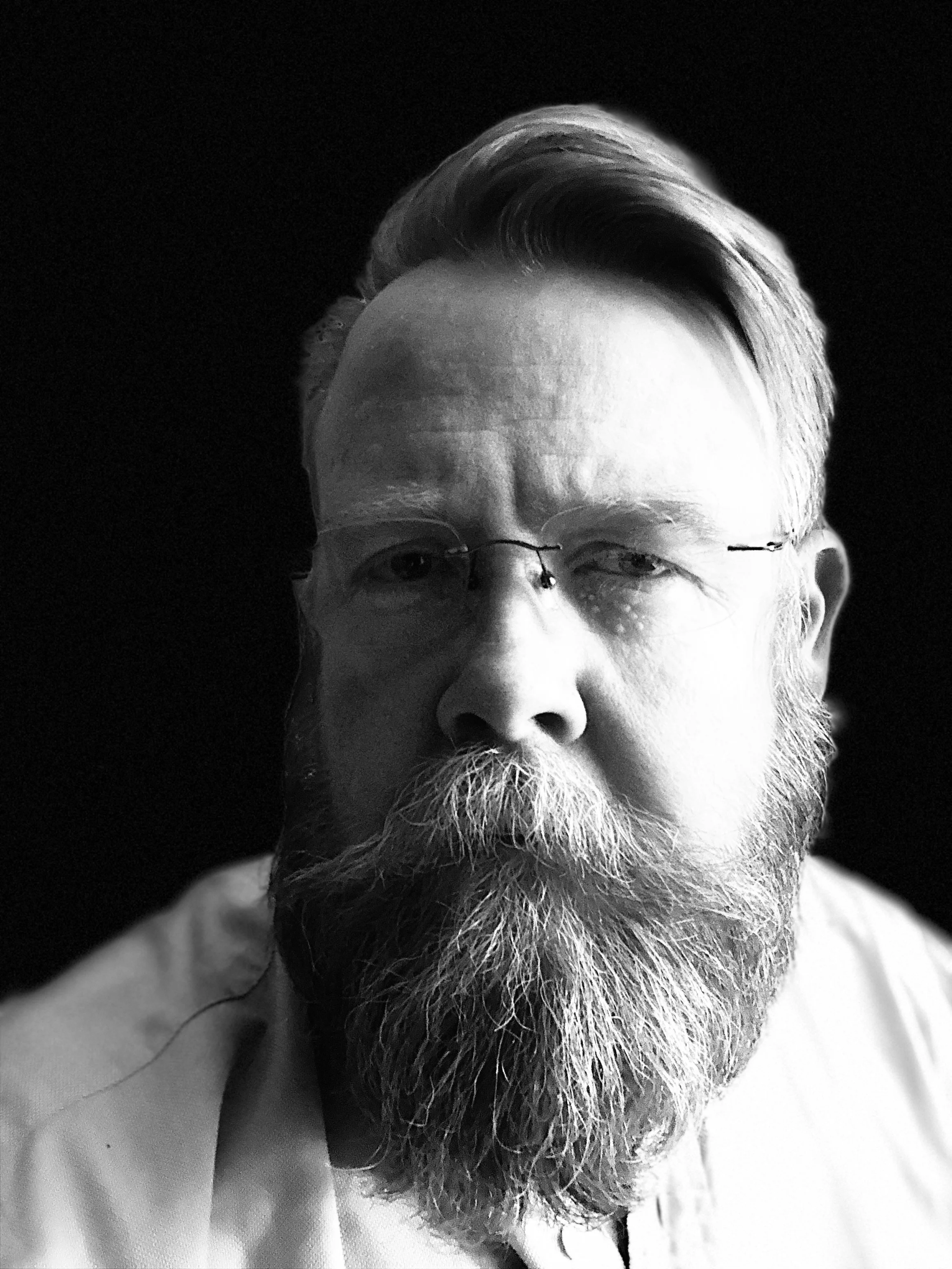 Black and white close-up portrait of an older man with a full beard and glasses, looking at the camera against a dark background.
