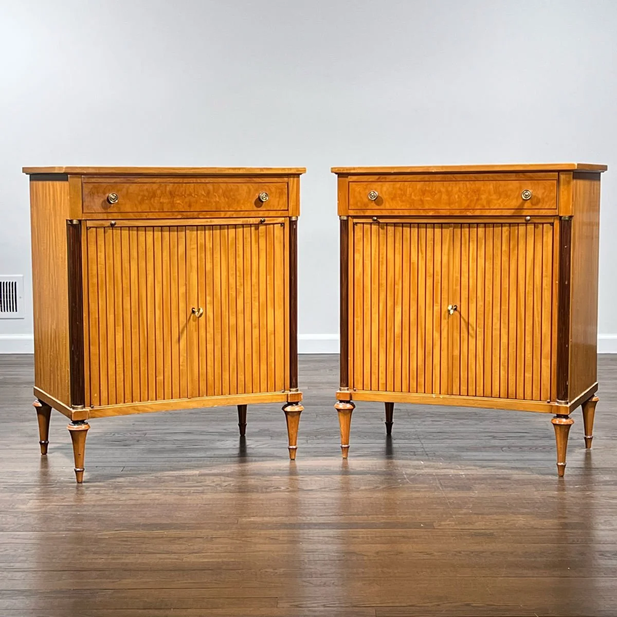 Two wooden cabinets with vertical slats on the doors and small round knobs, standing on tapered legs against a plain white wall.