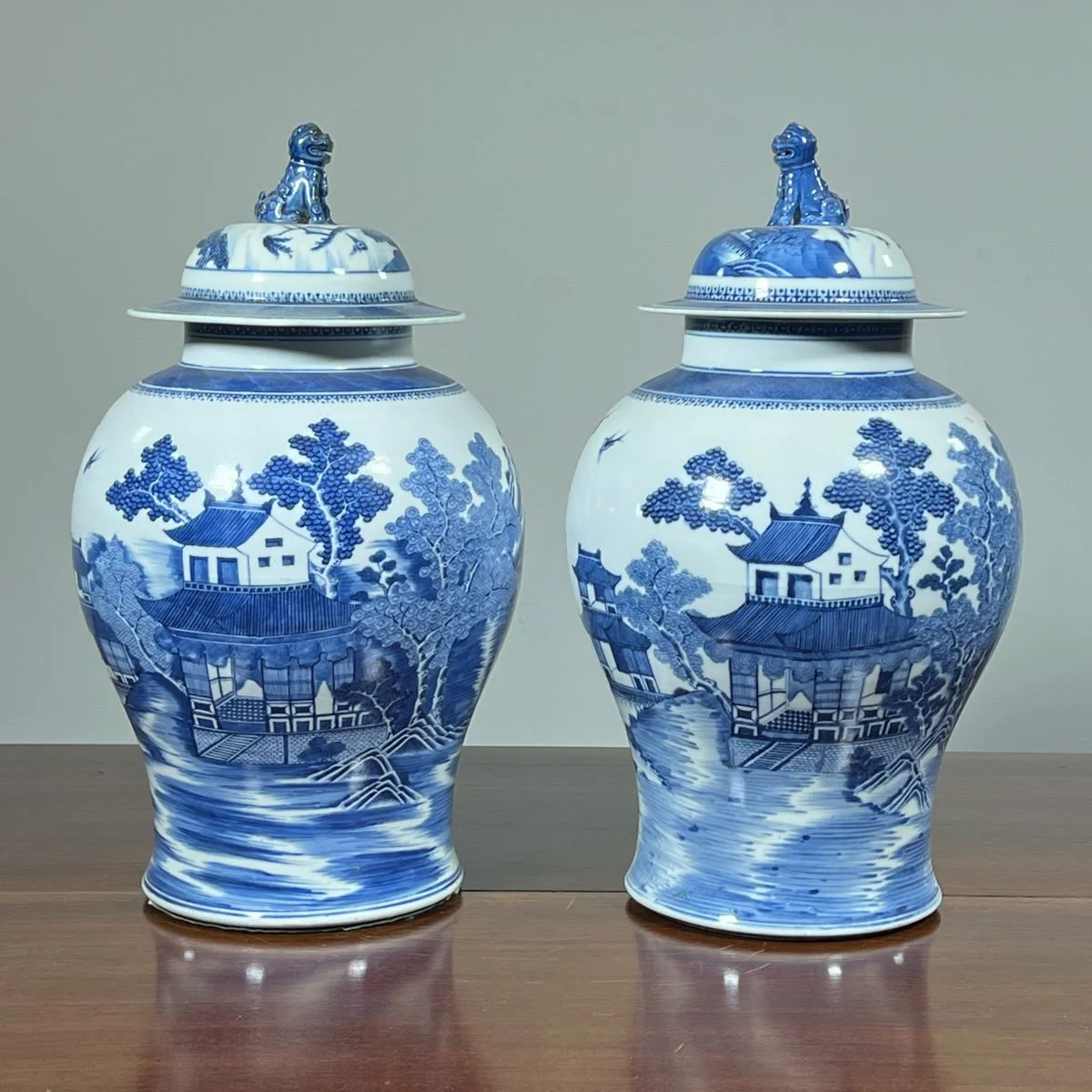 Pair of blue and white porcelain vases with lids, featuring landscape scenes with trees, buildings, and water, placed on a wooden surface.