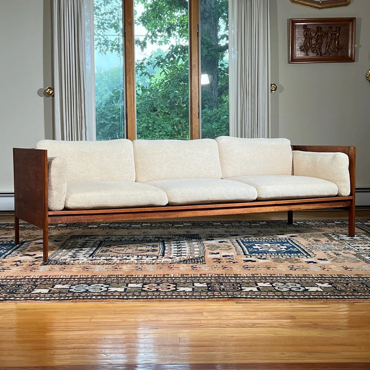 Living room scene with a cream-colored sofa with wooden frame, tall window with curtains, green trees outside, framed wall art, patterned area rug, and hardwood floor.