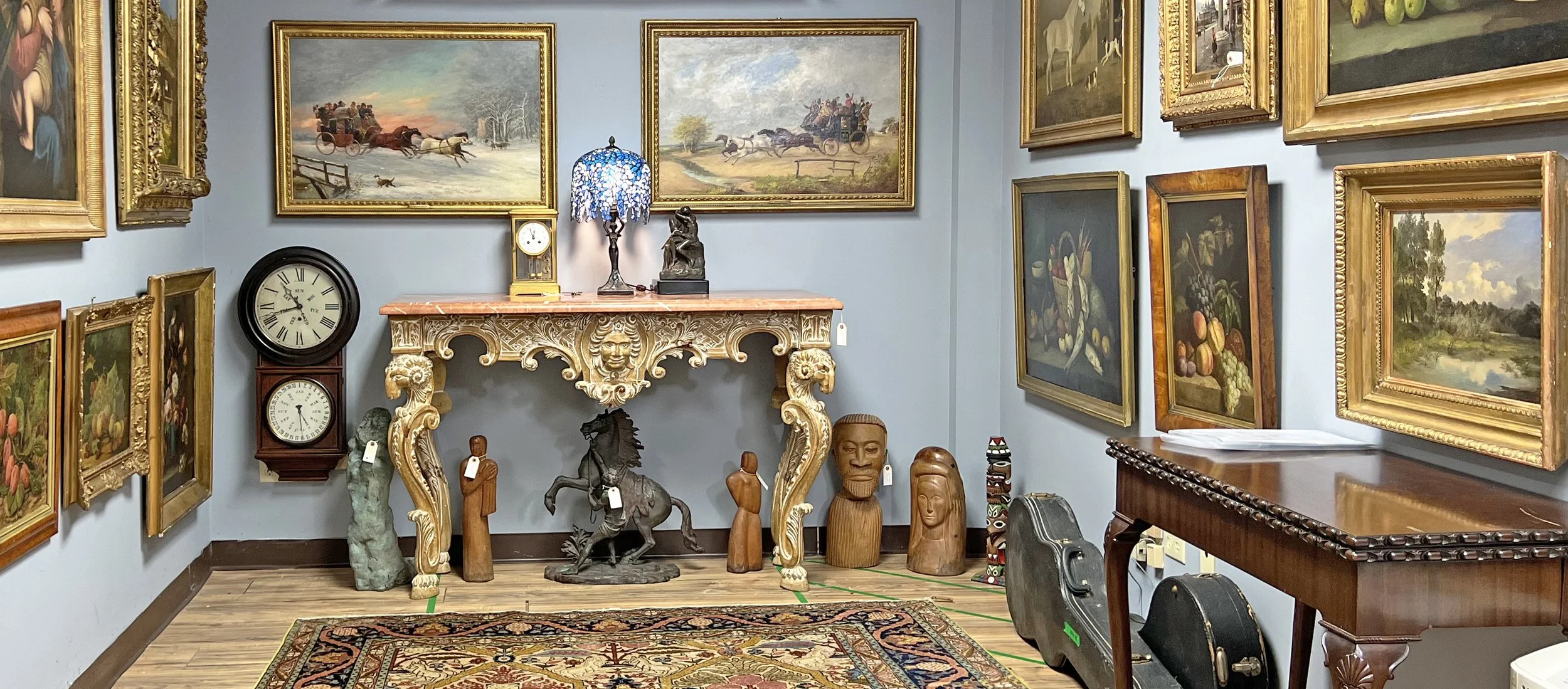An art gallery showcasing framed landscape and still life paintings on light blue walls, with a decorative ornate fireplace mantel, statues, and vintage clocks on the floor. There's a wooden table and a patterned rug on the wooden floor.