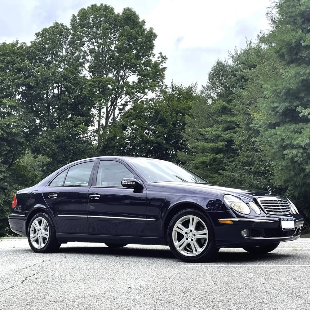 A black Mercedes-Benz sedan parked on a paved surface with green trees and a partly cloudy sky in the background.
