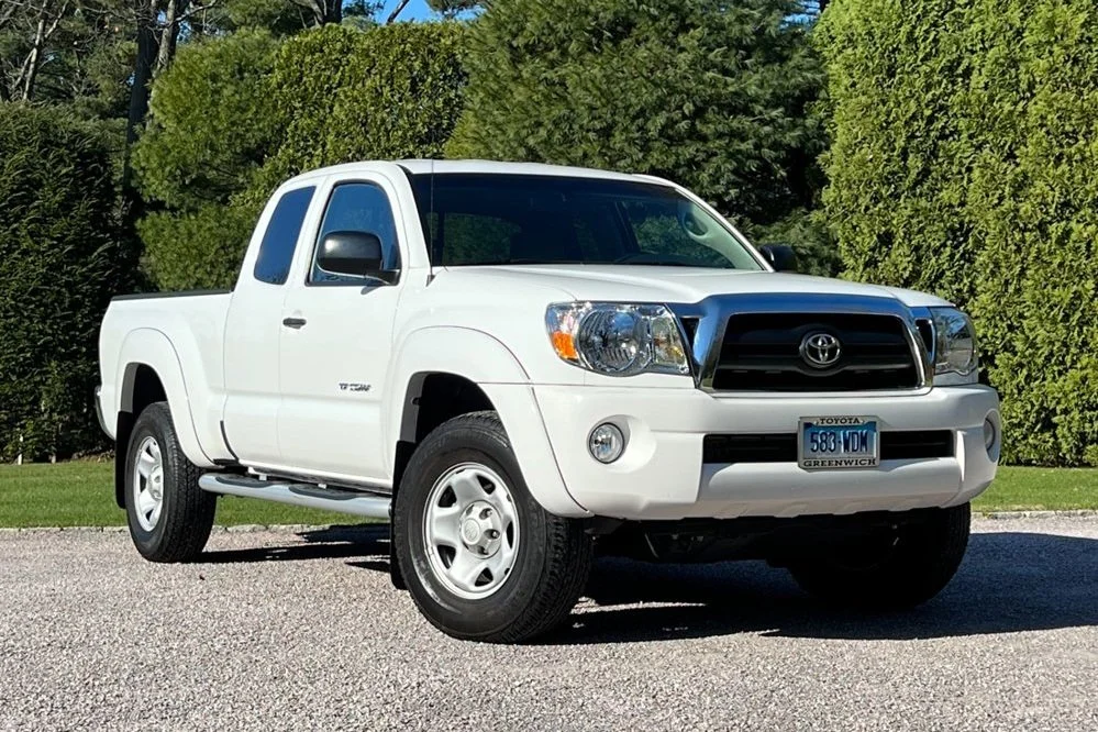 White Toyota Tacoma pickup truck parked on gravel with green bushes and trees in the background.