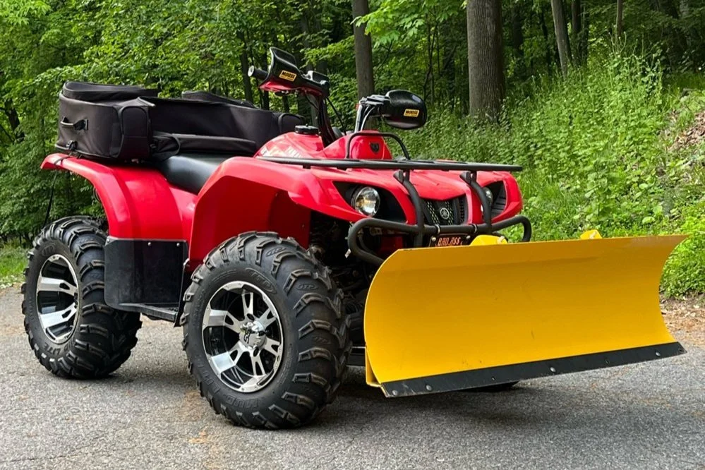 A red all-terrain vehicle (ATV) equipped with a yellow snow plow attachment on the front, parked on a paved road with green trees and grass in the background.