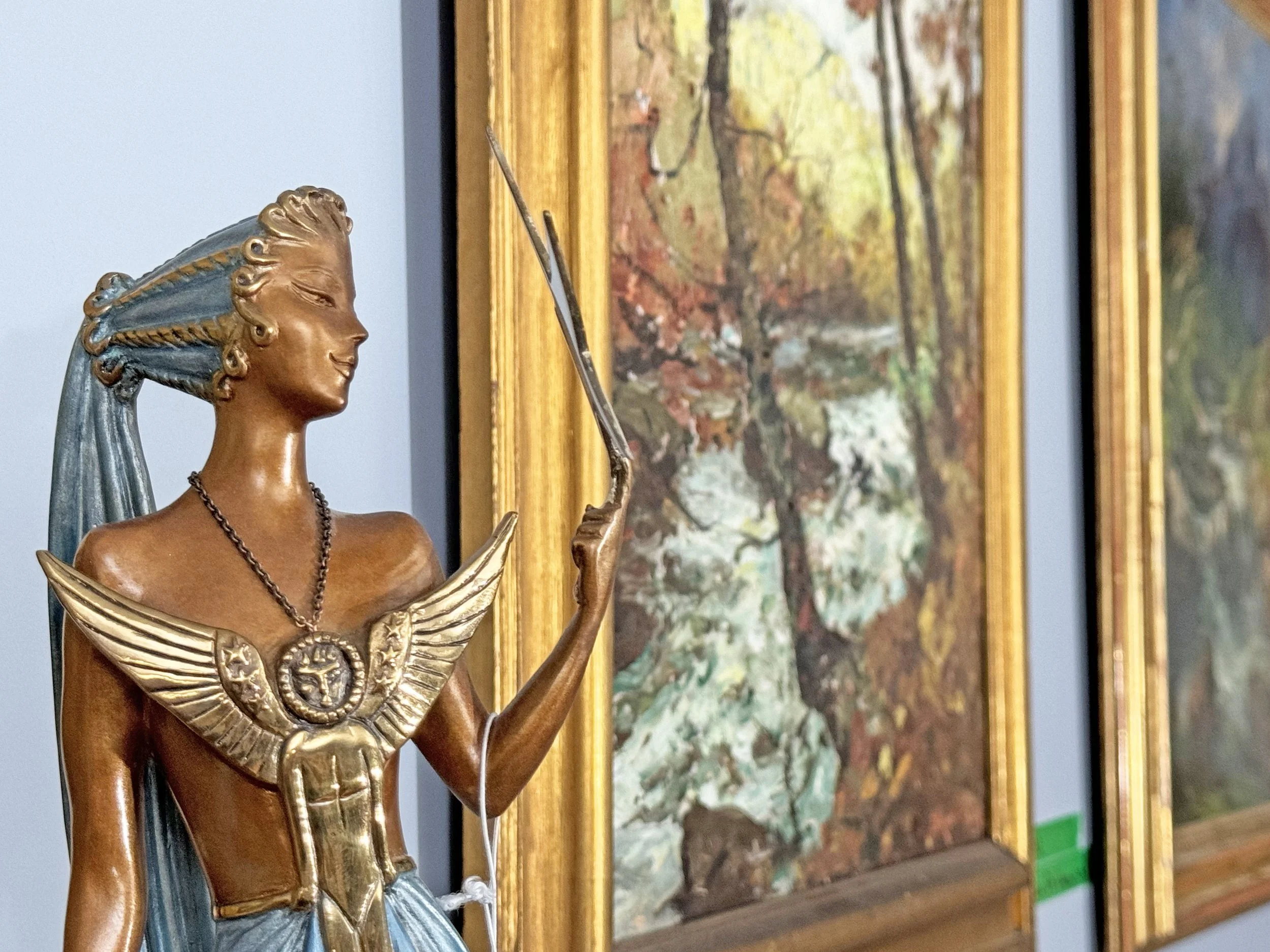 Bronze Erte statue of a woman holding a pointer, dressed in a gown with wings and a medallion necklace, in an art gallery with framed landscape paintings, auctioned and photographed by ACES Gallery.