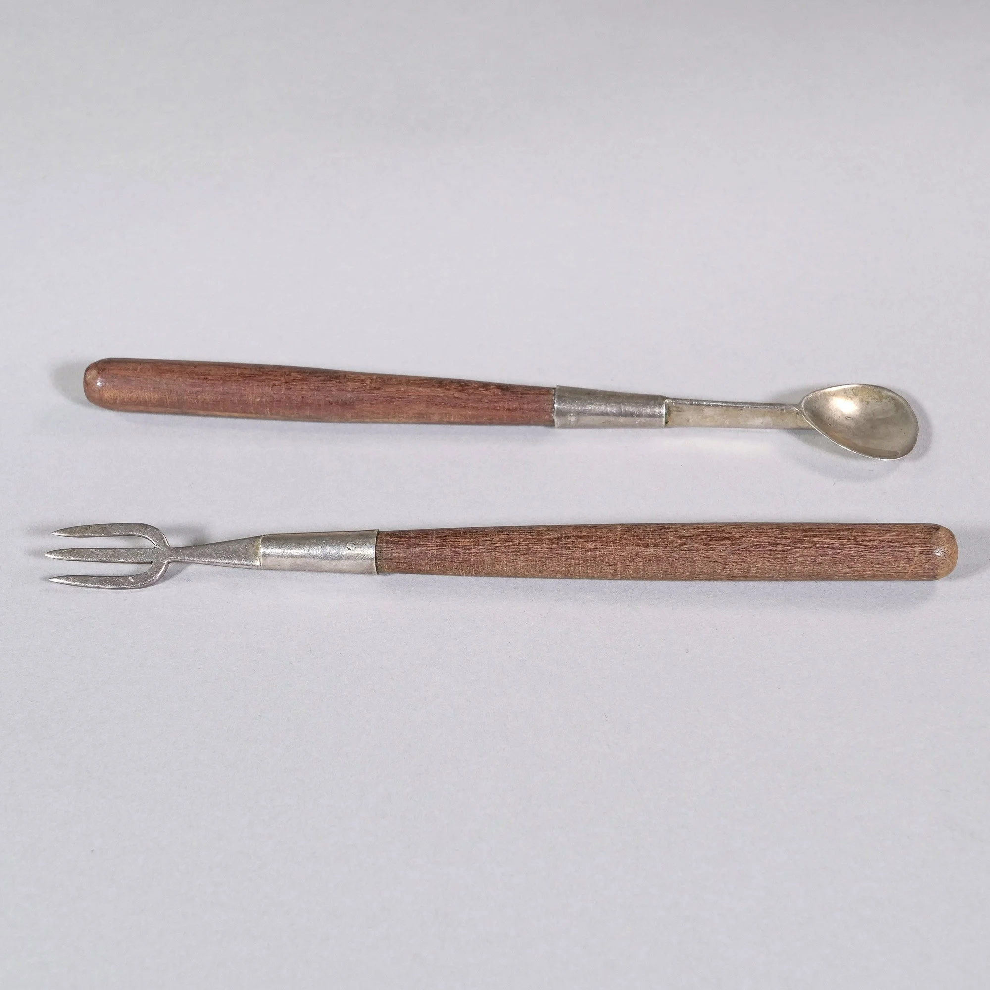 Two vintage silver serving tools with wooden handles, one with a small spoon and the other with a forked end, laid on a white background, auctioned and photographed by ACES Gallery.