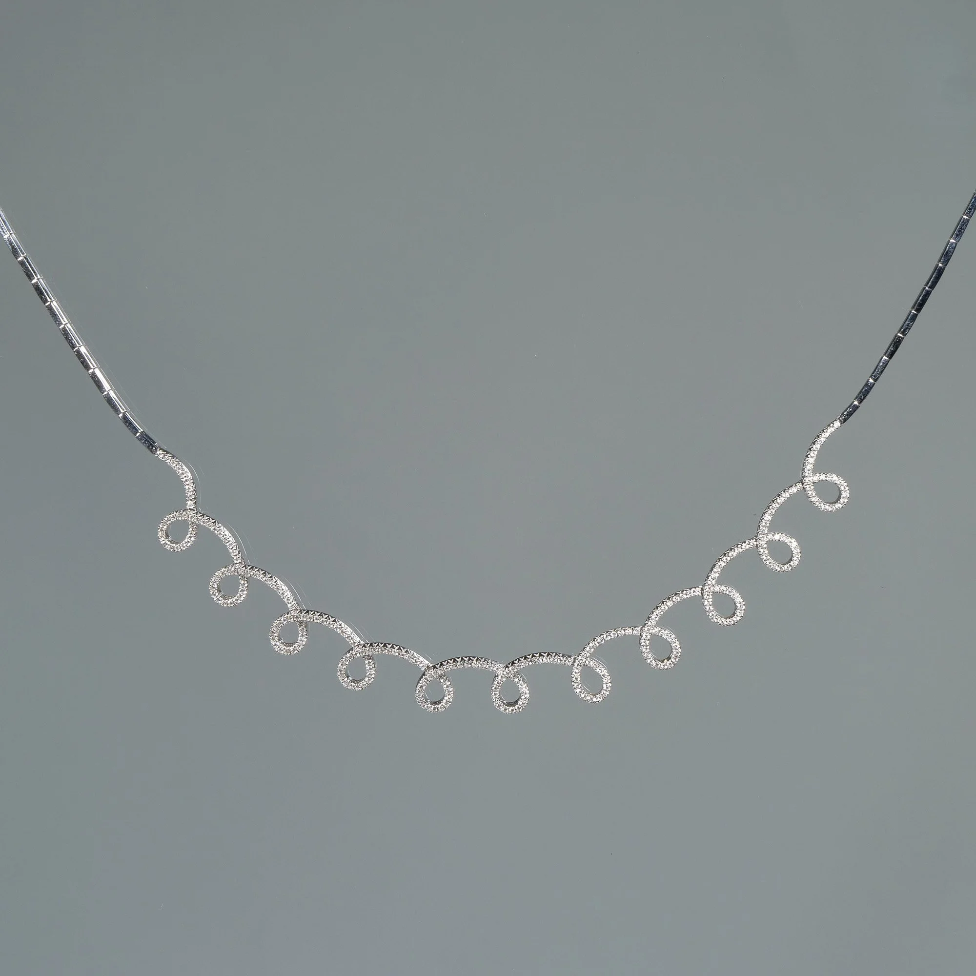 A white gold necklace with a decorative chain of loops and circles with melee diamonds, displayed against a gray background, auctioned and photographed by ACES Gallery.