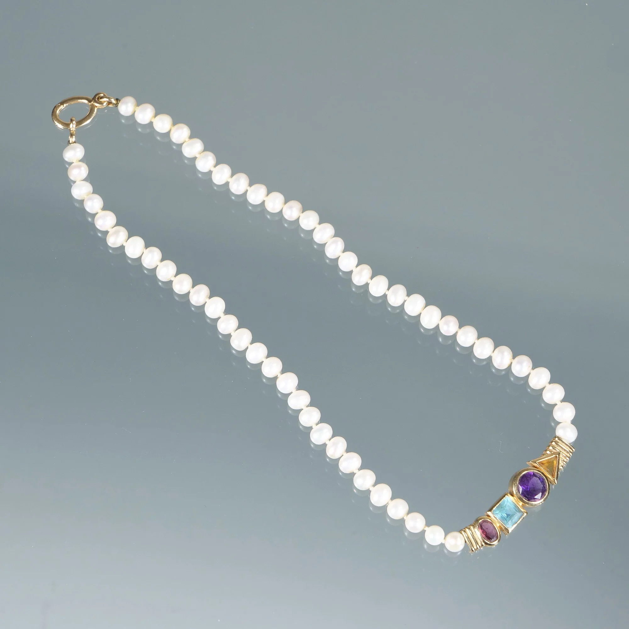 A pearl necklace with a gold clasp and colorful gemstone accents on a reflective surface, auctioned and photographed by ACES Gallery.
