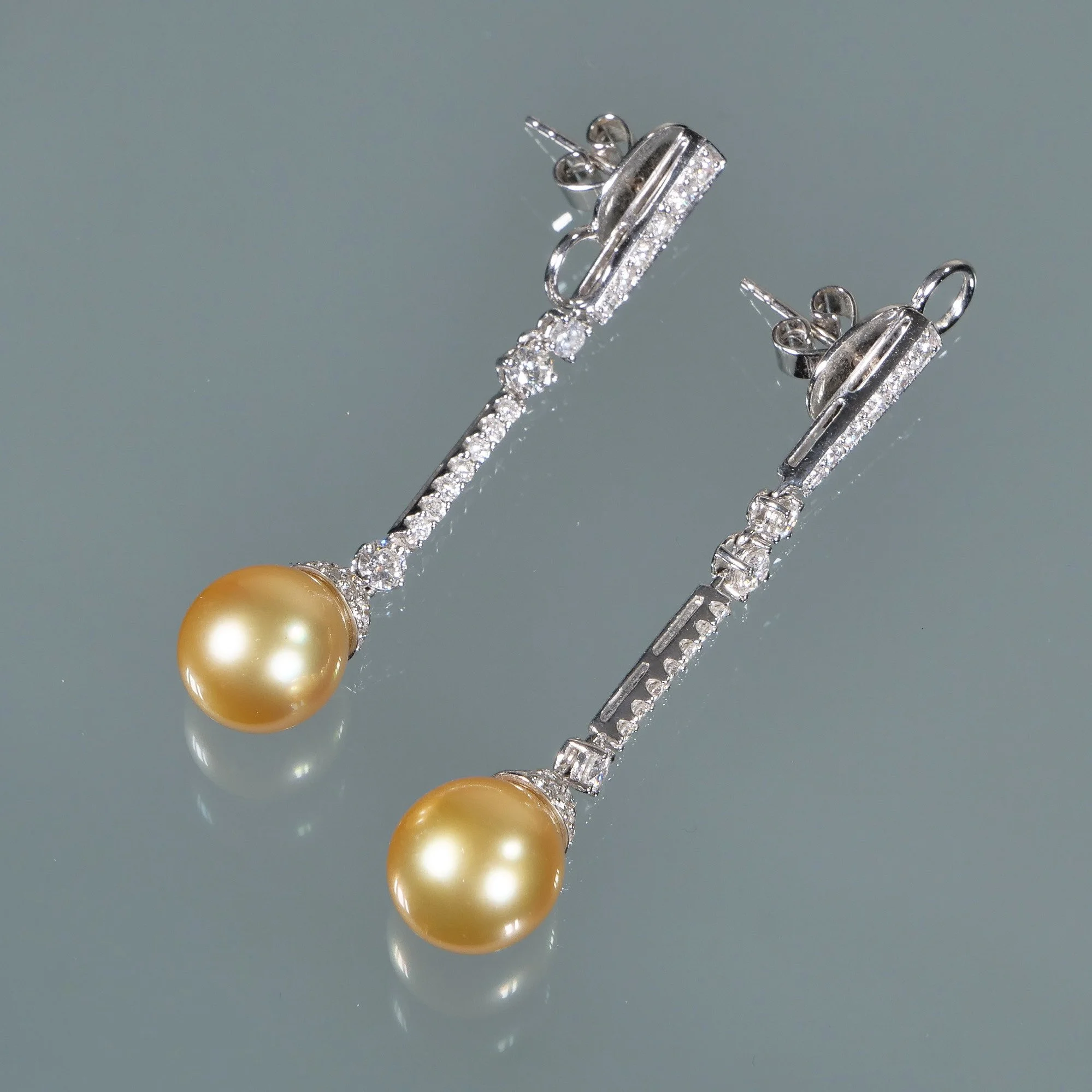 Pair of white gold earrings with yellow pearls and embedded diamonds, laid on a reflective surface, auctioned and photographed by ACES Gallery.