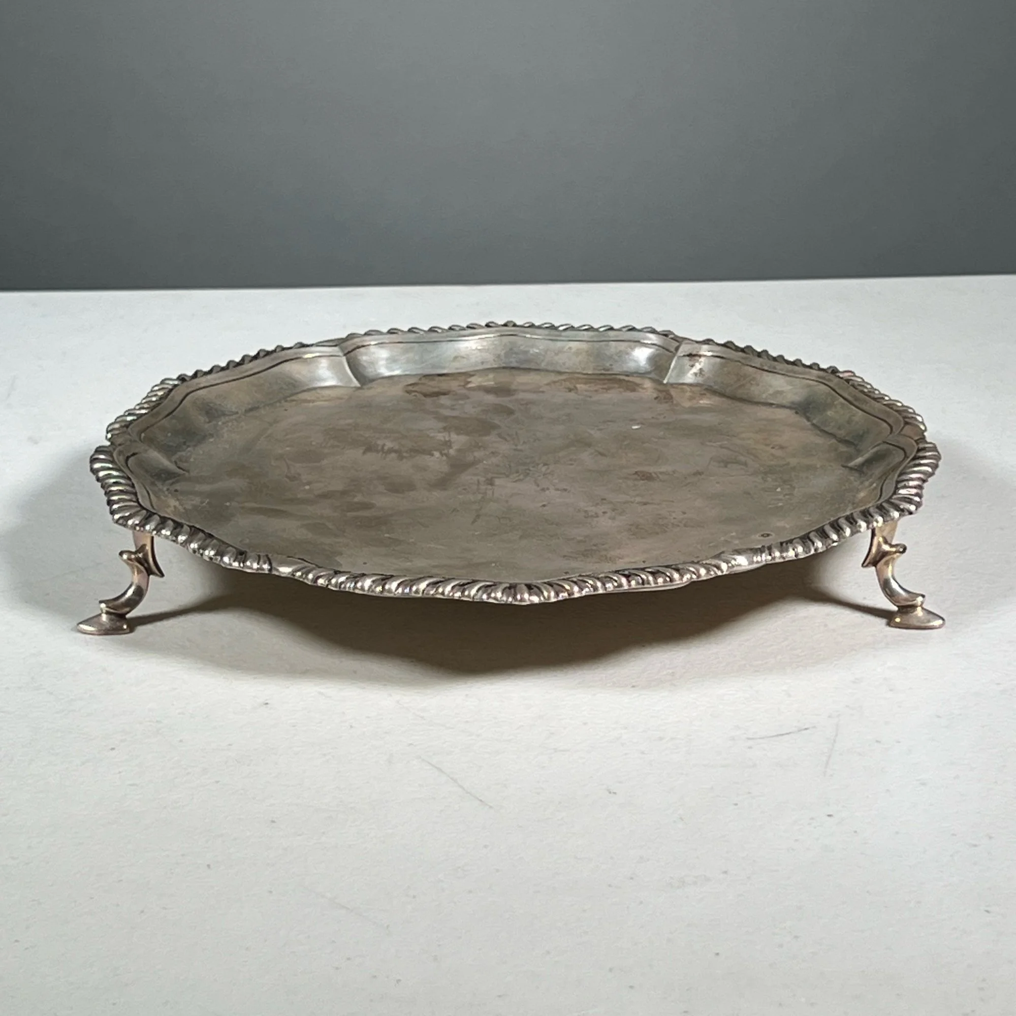 Silver decorative tray with ornate edges and small curved feet, placed on a white surface against a gray background, auctioned and photographed by ACES Gallery.