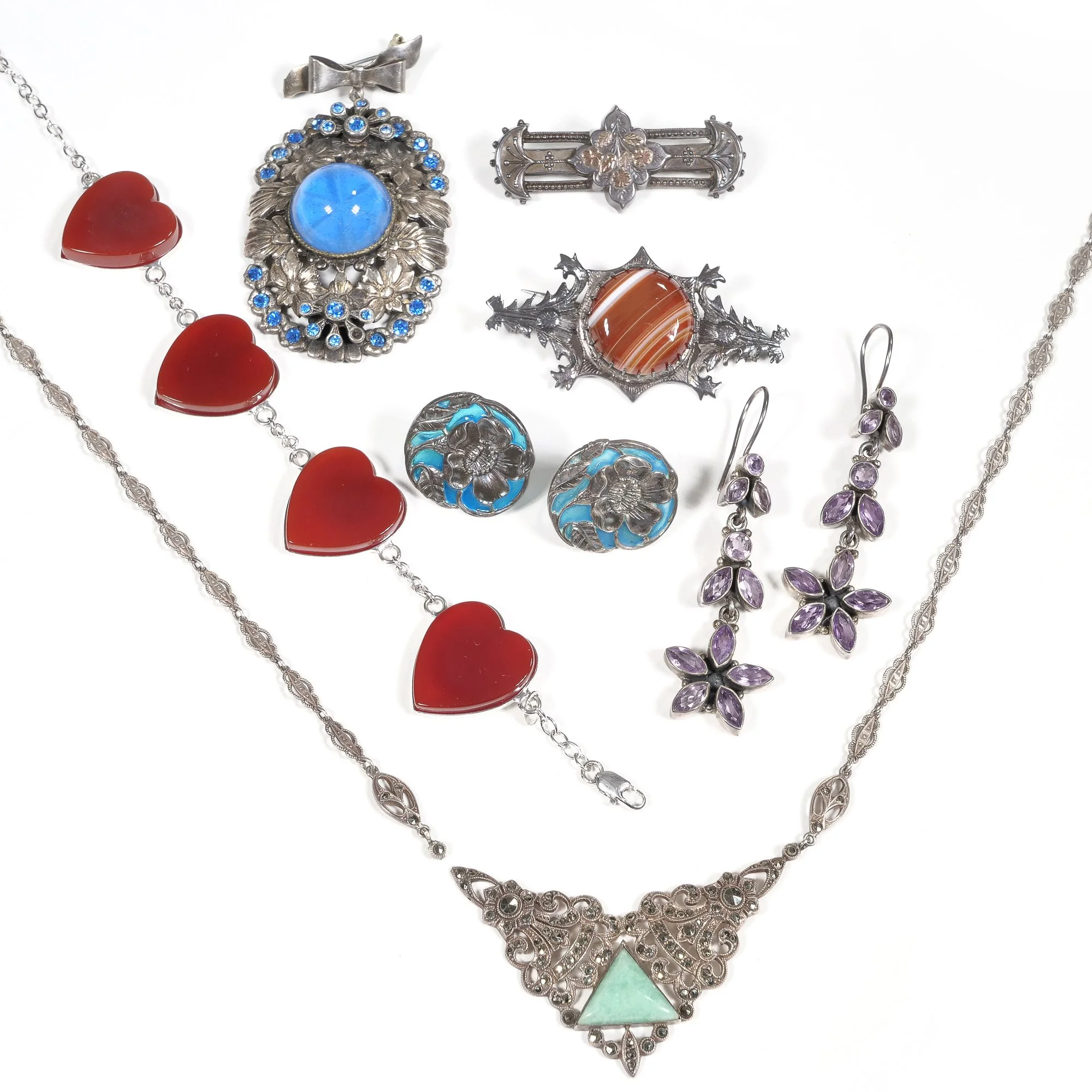 Collection of vintage jewelry including a silver necklace with green triangle pendant, red heart-shaped bracelet, blue gemstone brooch, floral brooch, two small blue and silver earrings, and purple gemstone earrings, photographed by ACES.