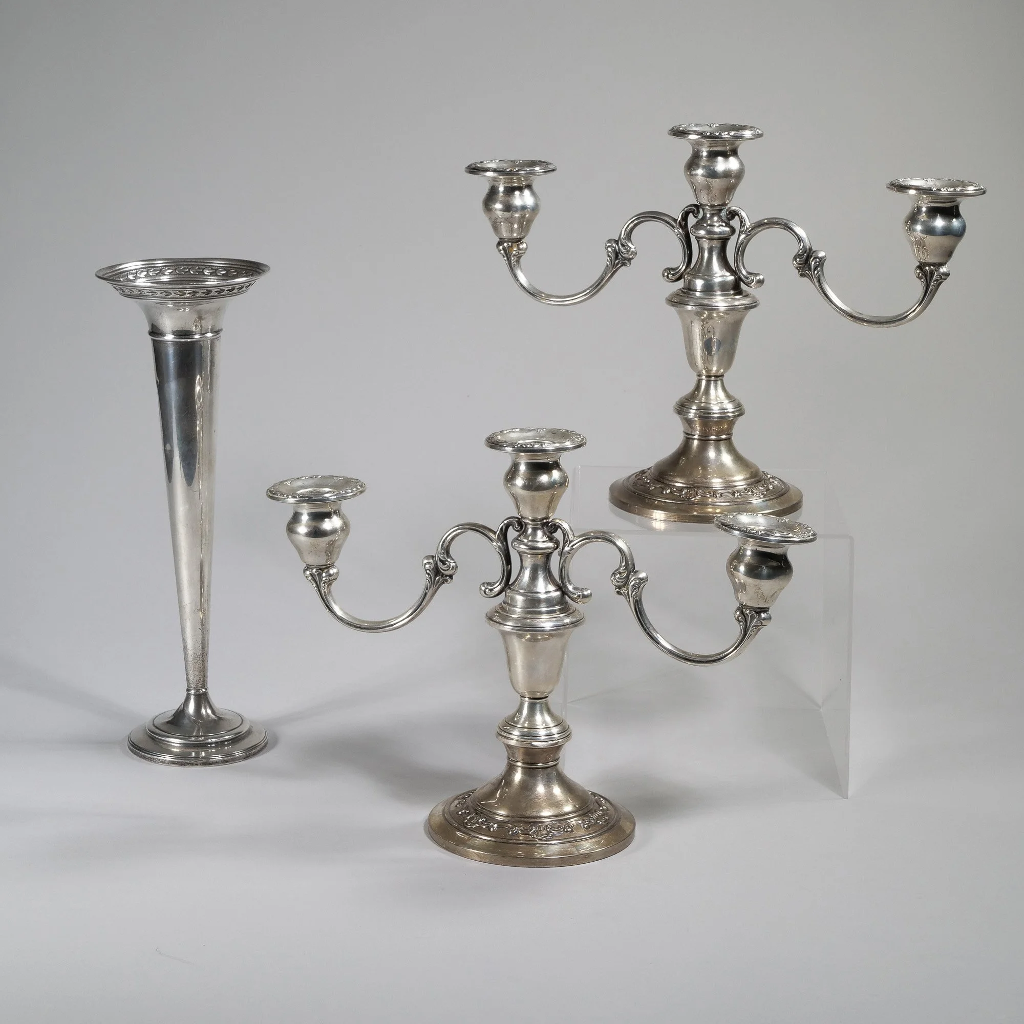 Three silver candle holders, two with three arms and one with a single tall stand, are displayed against a plain background, auctioned and photographed by ACES Gallery.