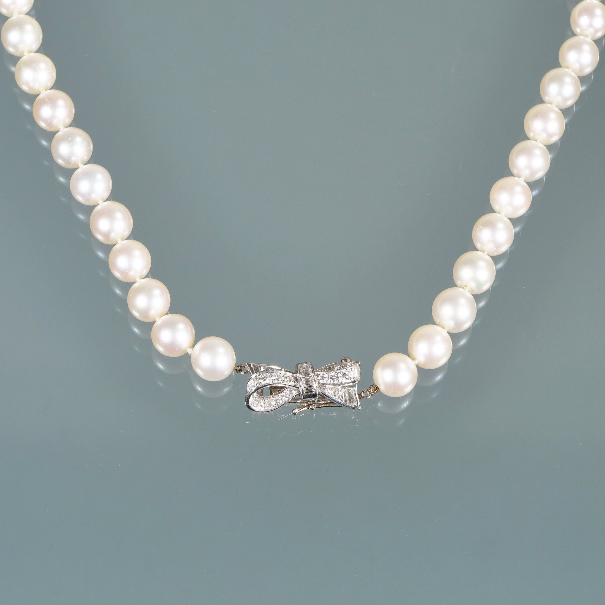 A string of white pearls with a silver bow-shaped clasp that has embedded diamonds on a gray background, auctioned and photographed by ACES Gallery.