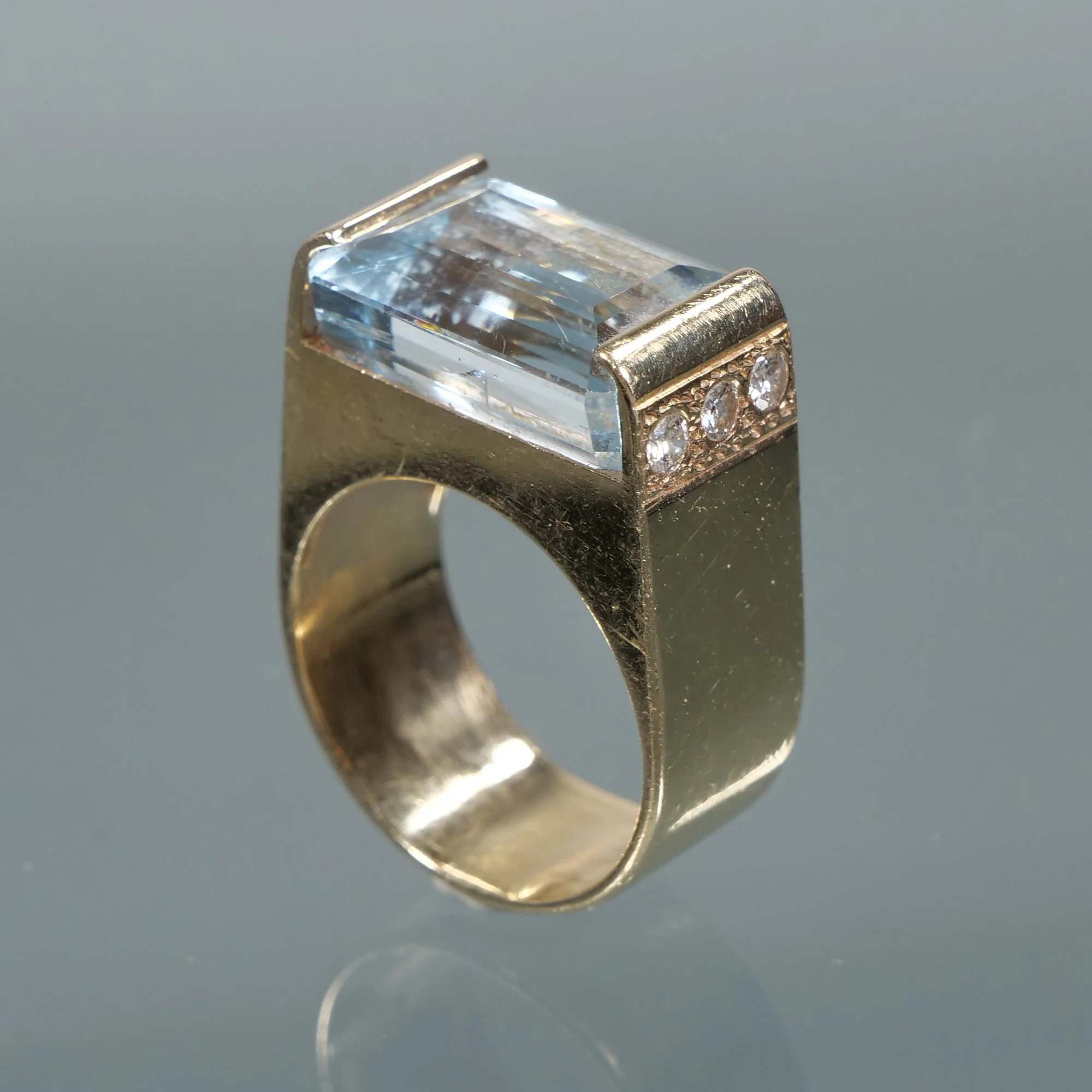 A gold ring with a large rectangular blue gemstone and smaller clear gemstones on the band, auctioned and photographed by ACES Gallery.