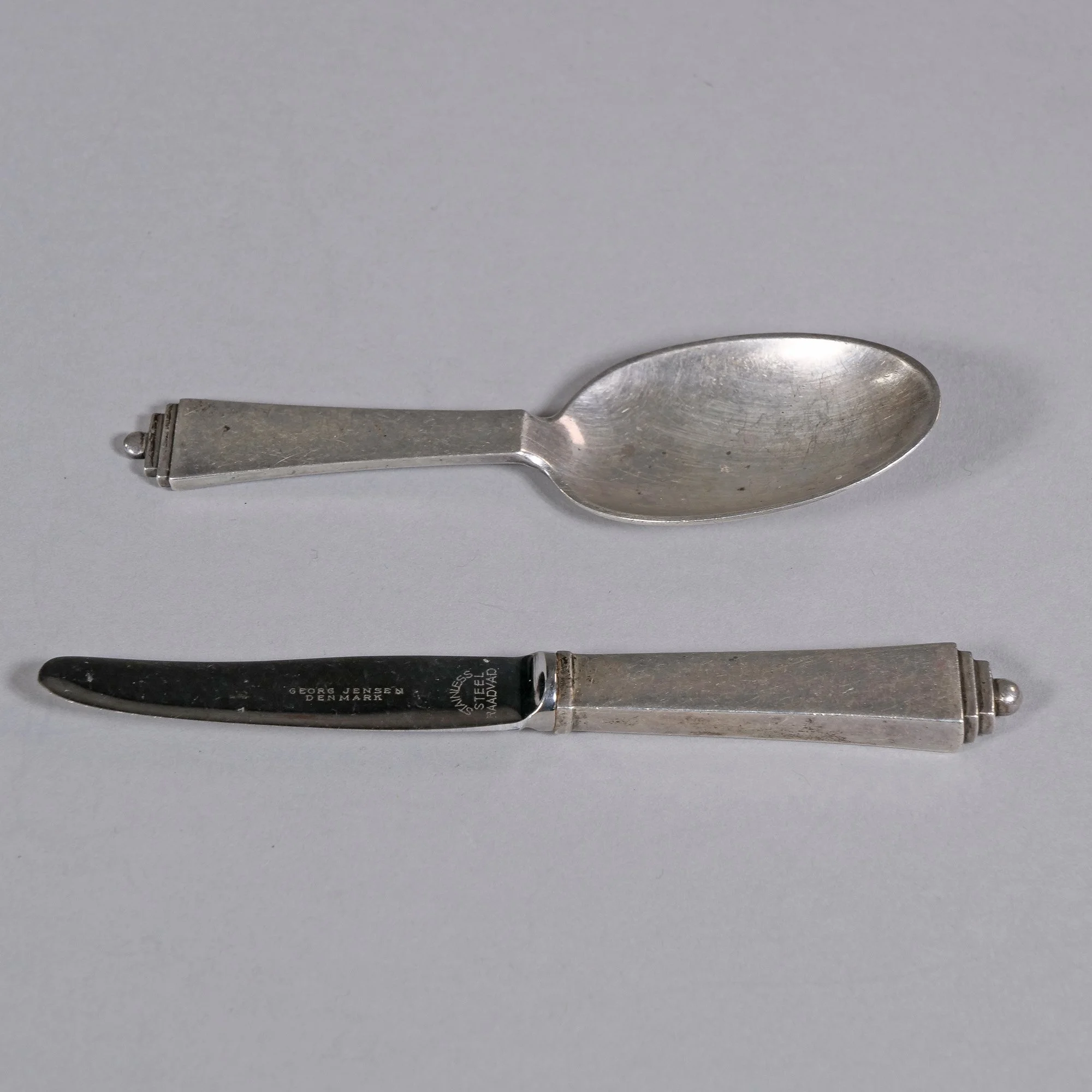 An antique silver spoon and a vintage silver knife laid on a plain surface, auctioned and photographed by ACES Gallery.