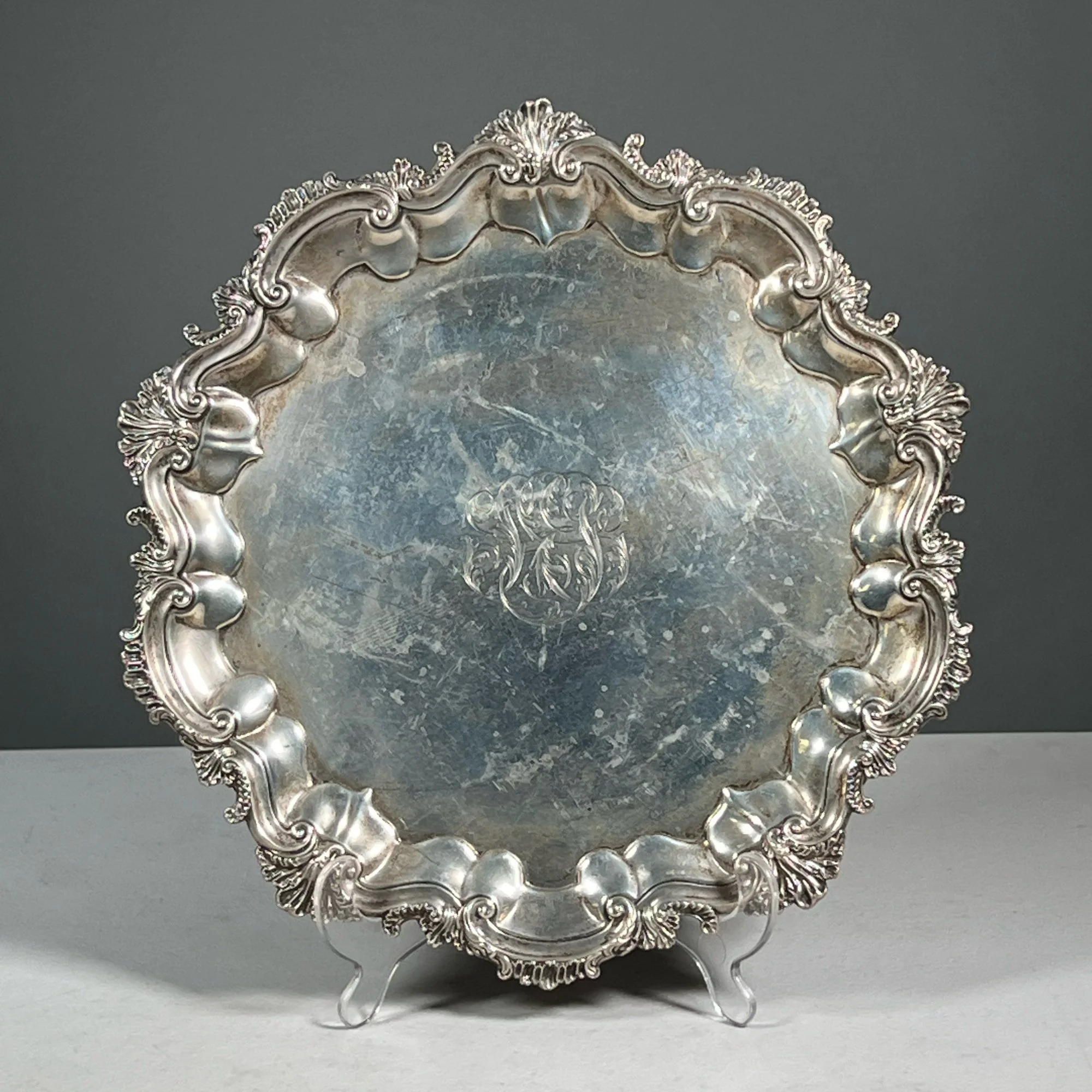 An ornate silver tray with intricate decorative edges and a monogram engraved in the center, placed on a white surface against a dark background, auctioned and photographed by ACES Gallery.