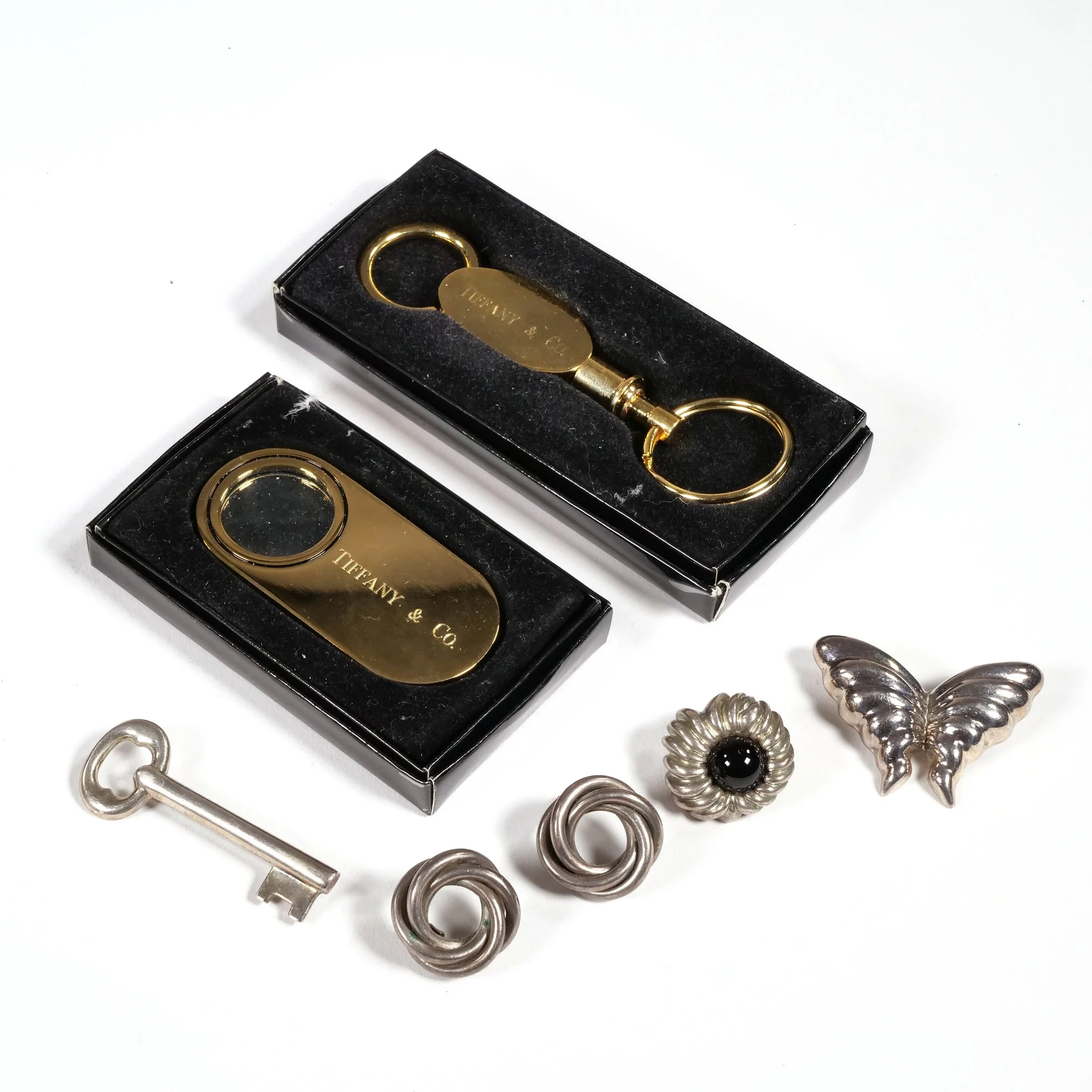 Two black jewelry boxes containing gold Tiffany & Co. keychains, surrounded by silver jewelry pieces including a key, a flower-shaped pin, a butterfly pin, and two spiral earrings, auctioned and photographed by ACES Gallery.