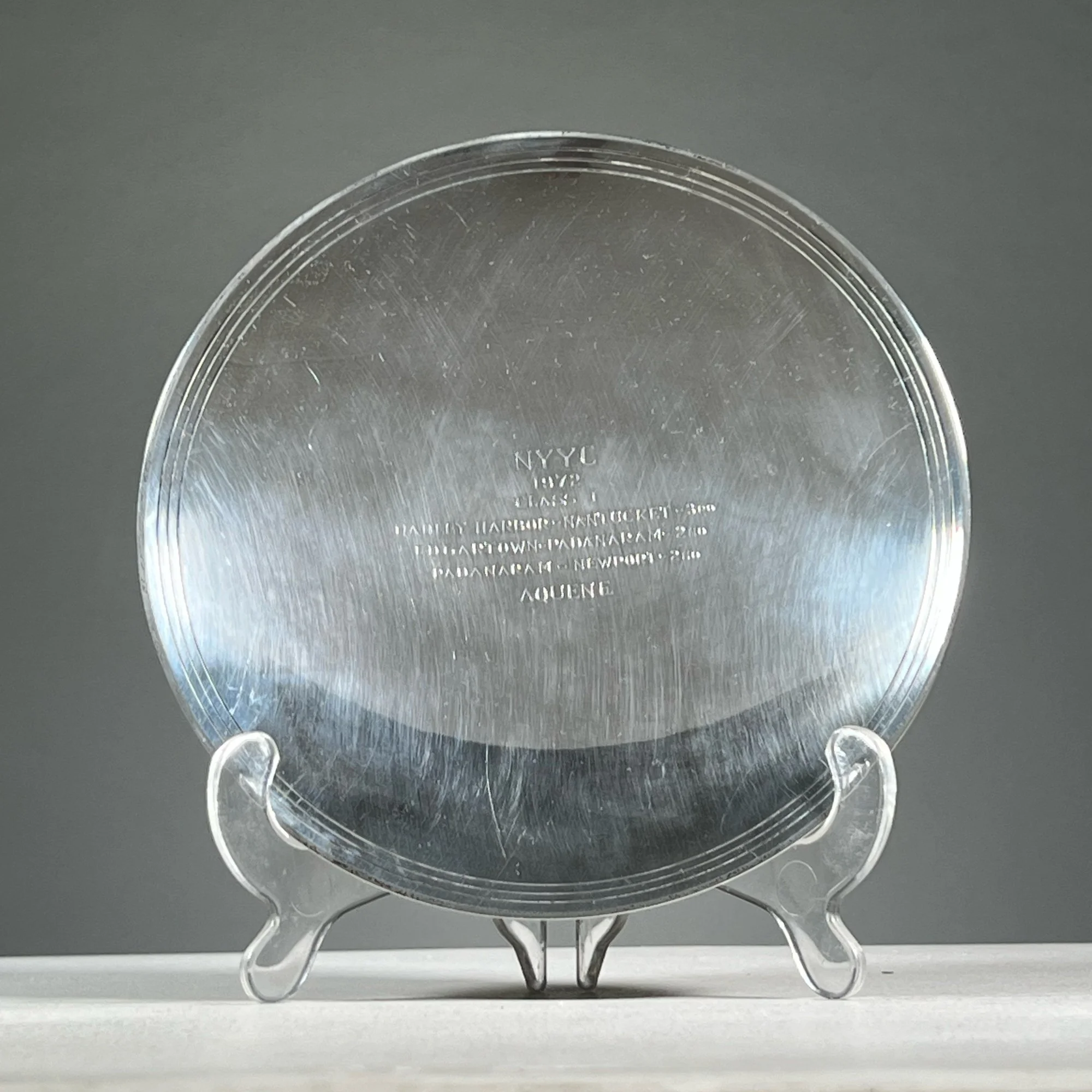 A sterling silver plate with text engraved on the back, displayed on a small stand against a plain gray background, auctioned and photographed by ACES Gallery.