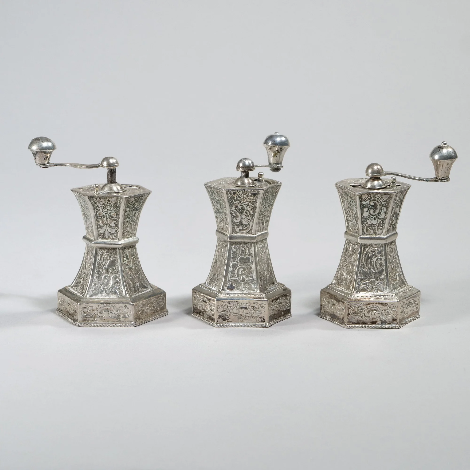 Three antique silver pepper mills with intricate engravings, lined up on a plain white background, auctioned and photographed by ACES Gallery.