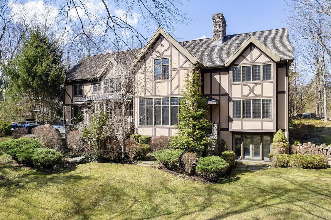 A large, two-story house with Tudor-style exterior, multiple windows, and a manicured lawn and garden in the front yard, under a partly cloudy sky, from an estate represented by ACES Gallery.