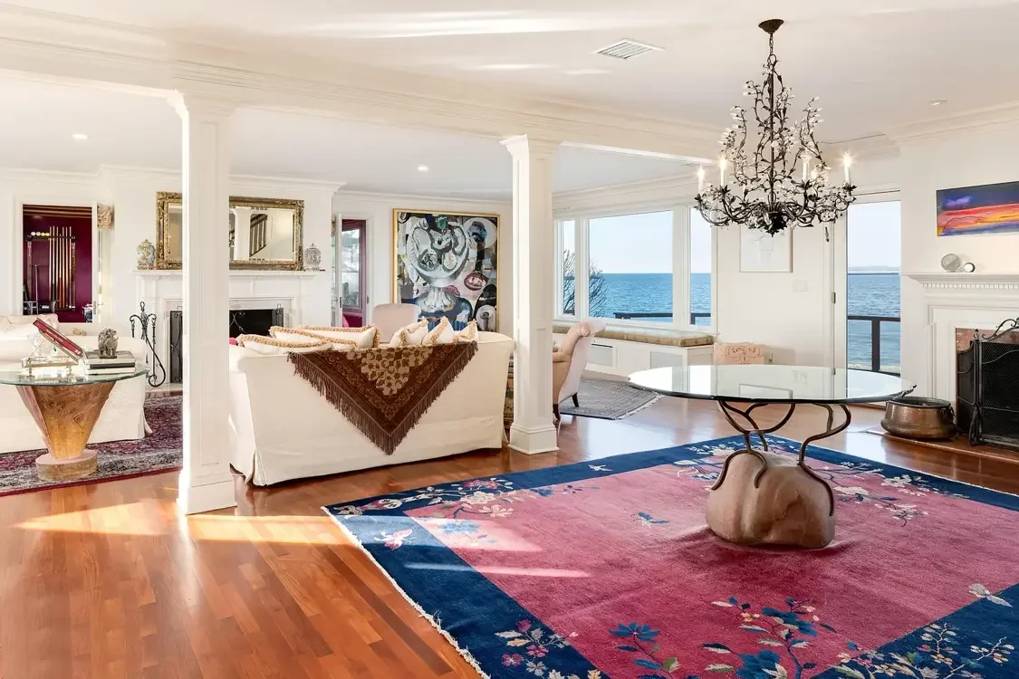 Bright living room with large windows overlooking the ocean, featuring a black chandelier, a pink and navy floral area rug, and various furniture including white sofas, armchairs, and a white fireplace, taken in ACES client's home, with auction items