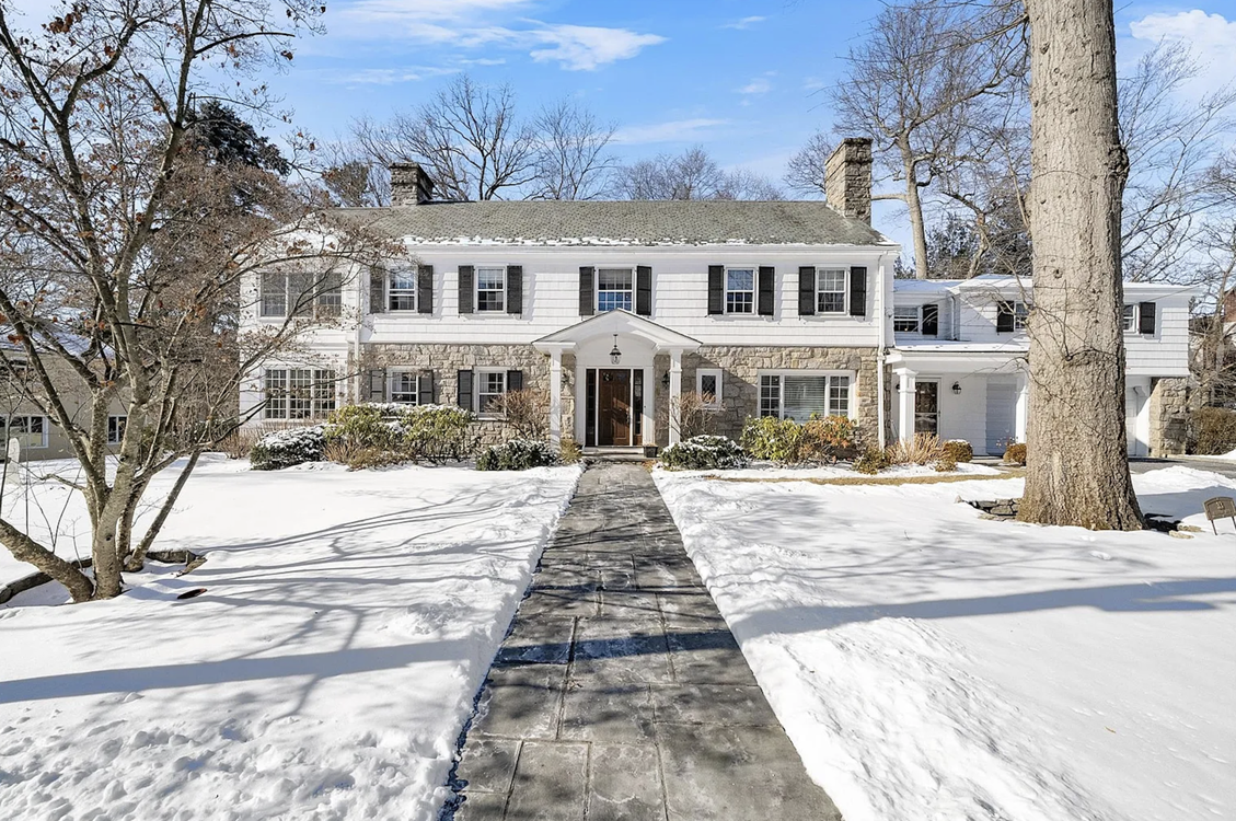 A large, white, two-story house with black shutters, a stone facade on the lower half, and a front porch with white columns, surrounded by snow-covered lawn and leafless trees, under a partly cloudy blue sky, representing an ACES Gallery estate.